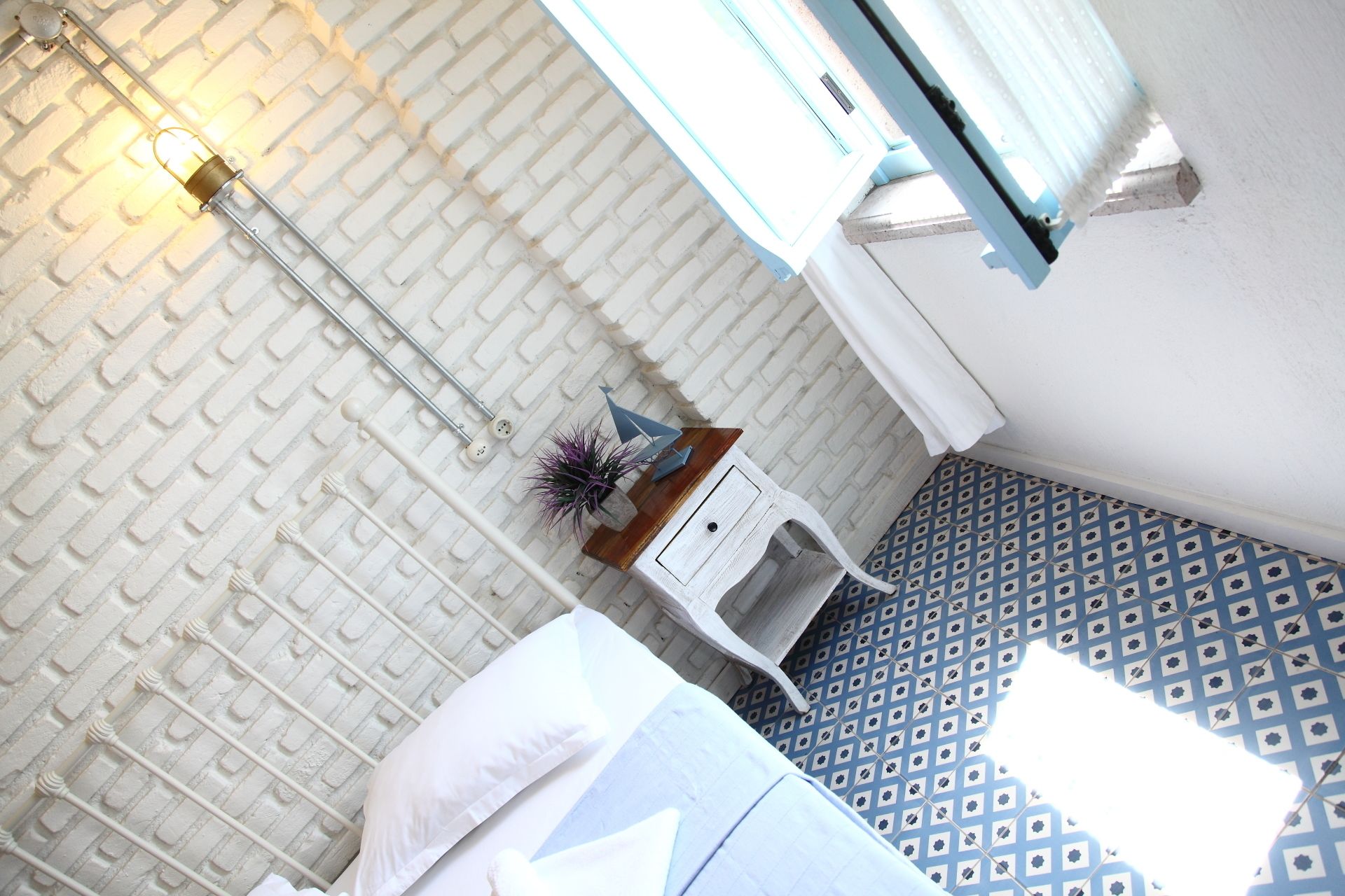 Een kamer met een witte bakstenen muur, een bed met witte lakens, een klein wit nachtkastje met een plant en een vloer met patroon.