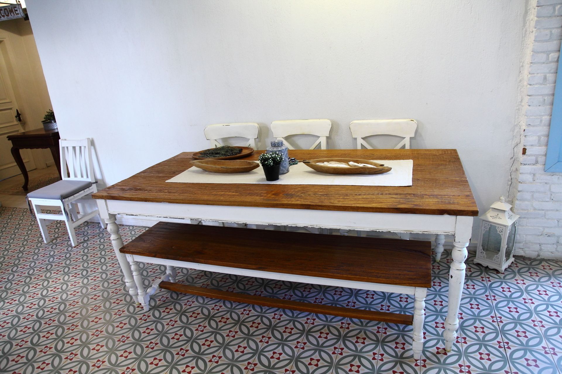 Una mesa con tapa de madera y patas blancas, acompañada de cuatro sillas blancas. Un estante pequeño está sujeto al frente de la mesa.