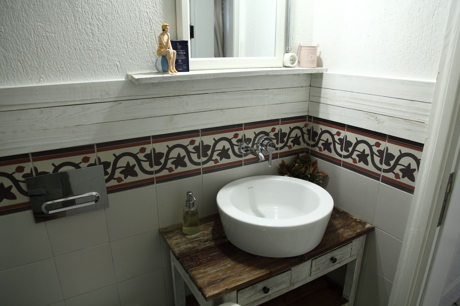 Un baño con lavamanos blanco, tocador de madera, azulejos decorativos y una ventana con repisa.