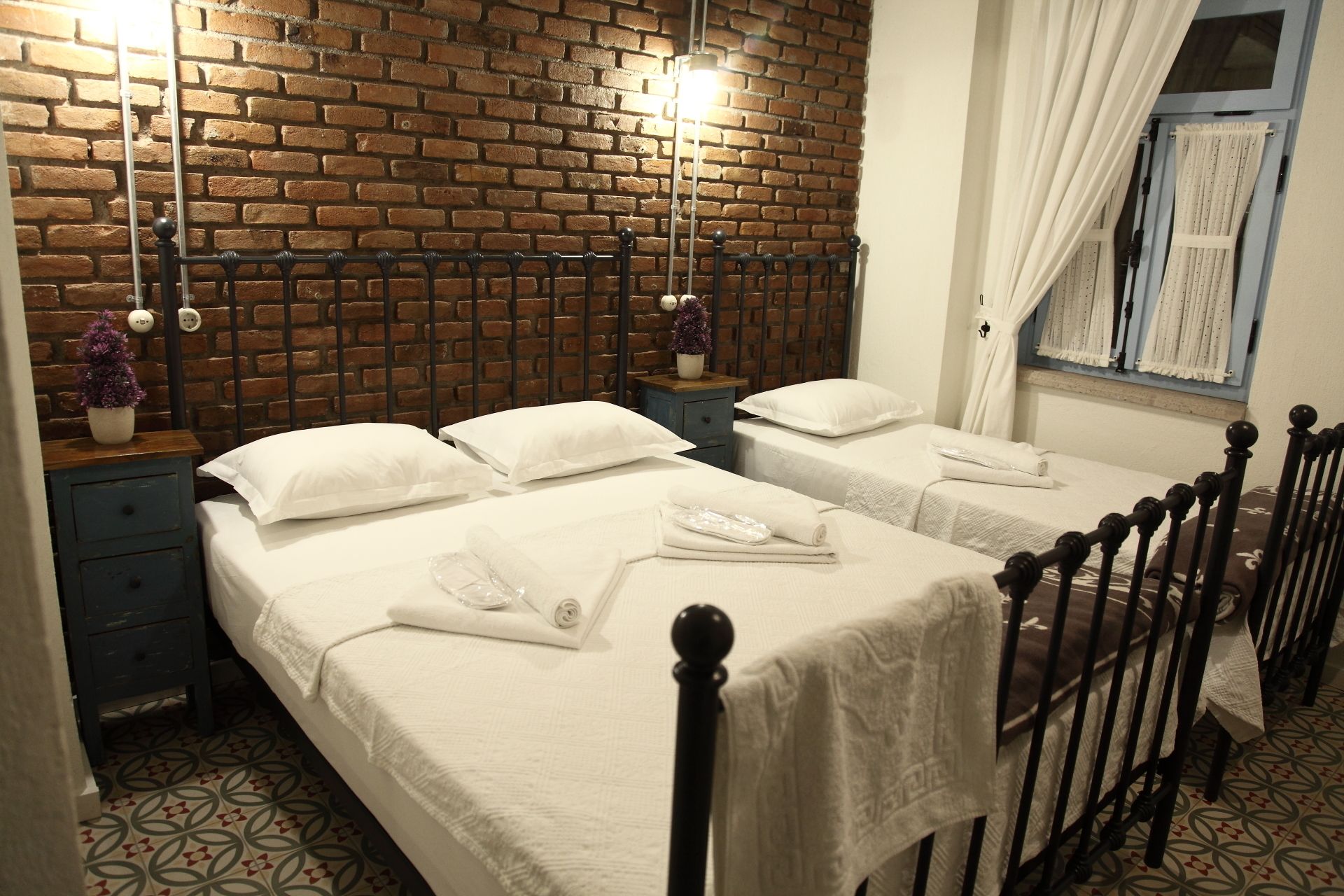 A hotel room with two beds, a brick wall, a window with curtains, and a small nightstand with a plant.