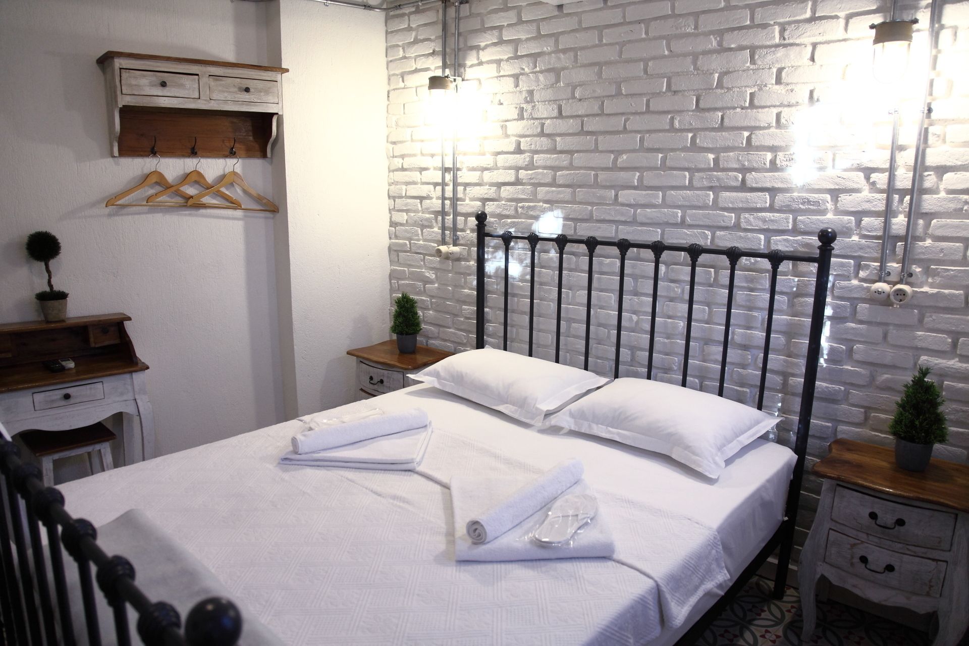 Cama bem arrumada com lençóis e almofadas brancos, mesa de cabeceira de madeira com uma pequena planta, e parede de tijolos brancos com cabeceira de metal.