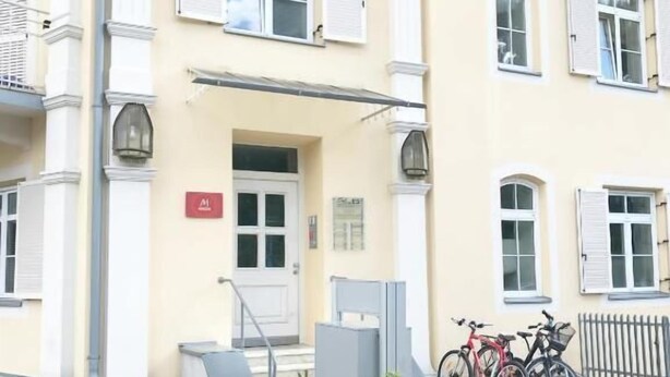 A building with a beige facade, white shutters, and a door with a red sign. There are two bicycles parked outside and a metal bench in front. - Manzoni Premium (Merano)