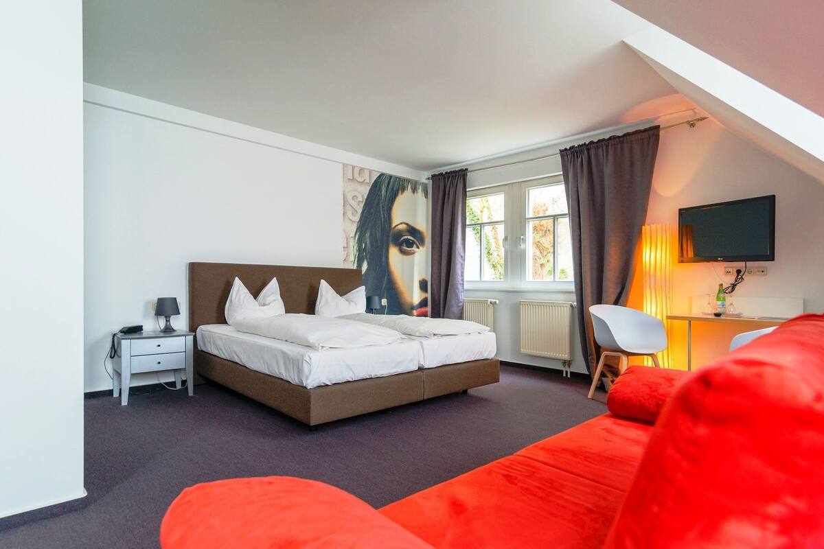 A bedroom with a bed, a desk with a chair, a TV, and a window with curtains.