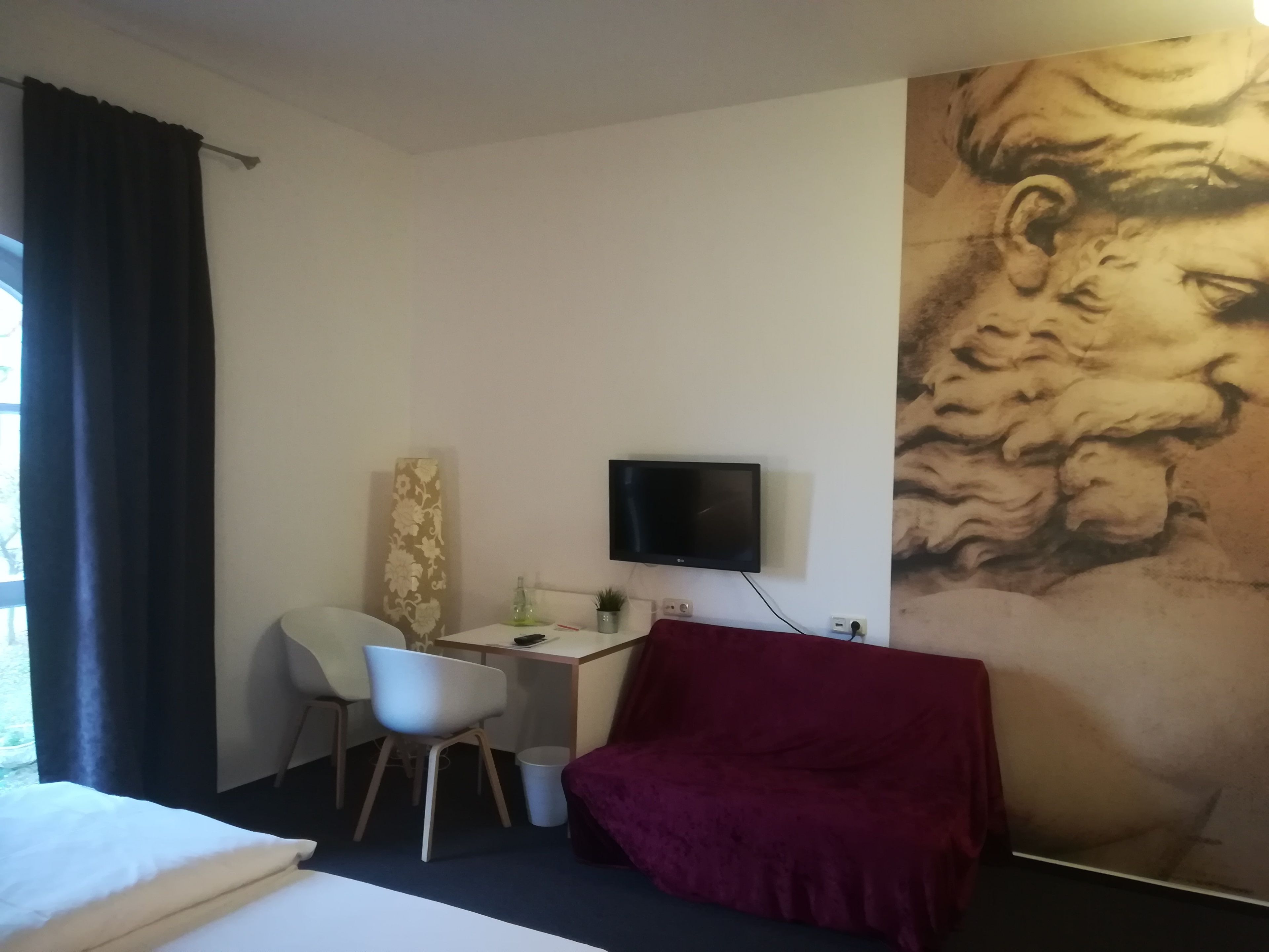 A hotel room with a bed, a desk with a chair, a television, and a large mural on the wall.