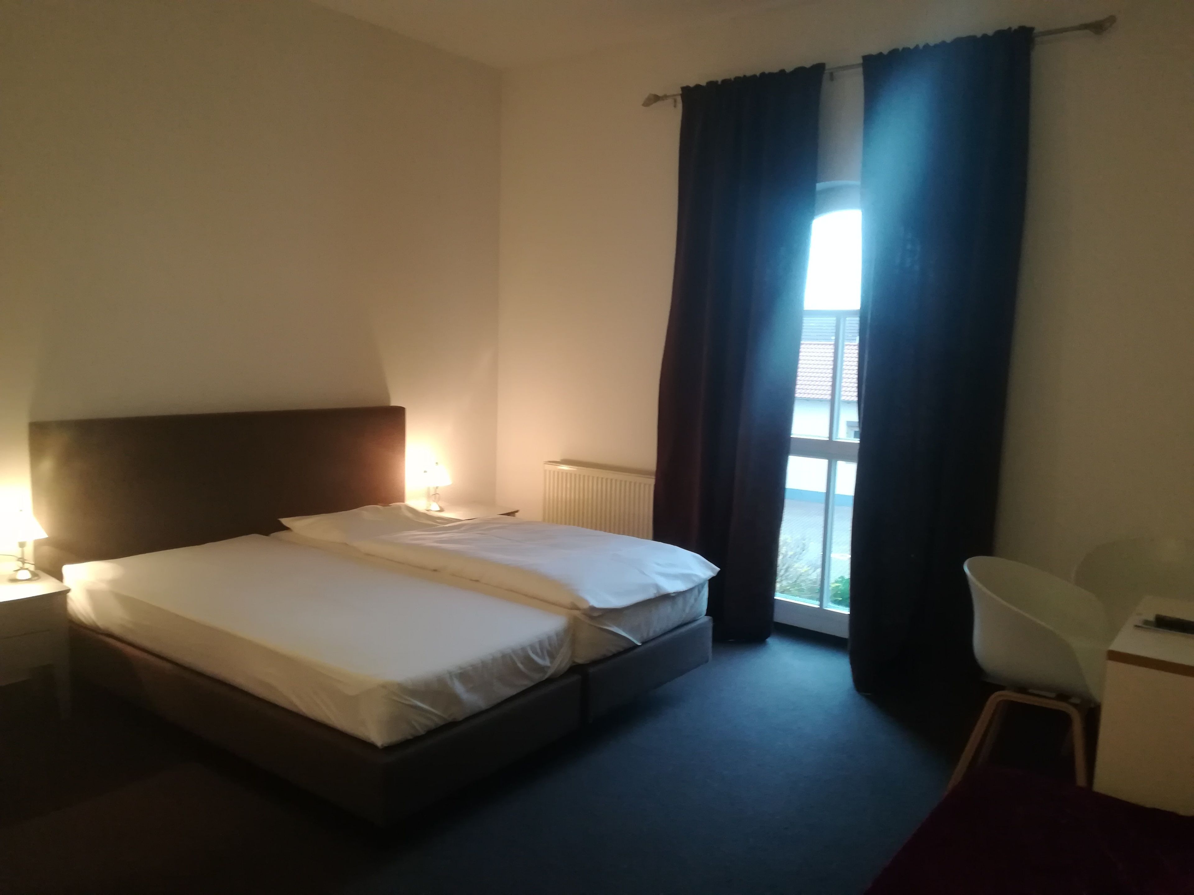 A hotel room with a bed, a nightstand, a lamp, a chair, and a window with curtains.