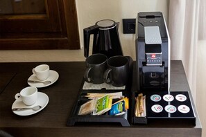 A coffee station with a coffee maker, two black mugs, two white cups, a tray with coffee packets, and a spoon. - Contessa Boutique Hotel (Chania)
