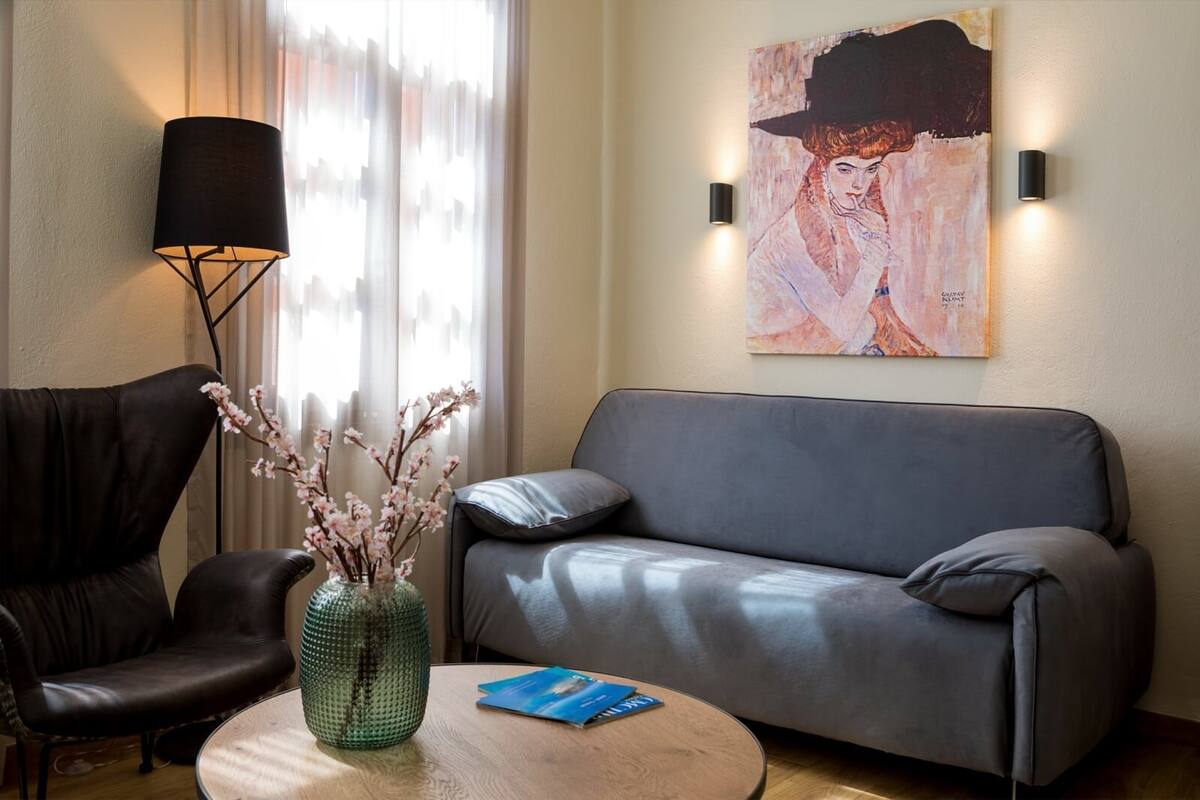 A modern living room with a leather sofa, a black armchair, a round wooden coffee table with a vase of flowers, and a painting on the wall.