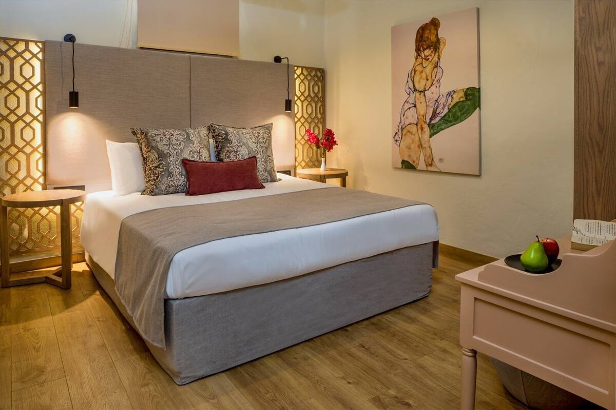 A modern hotel room with a large bed, a bedside table, a nightstand, a wall-mounted lamp, a painting, and a small desk with fruit.