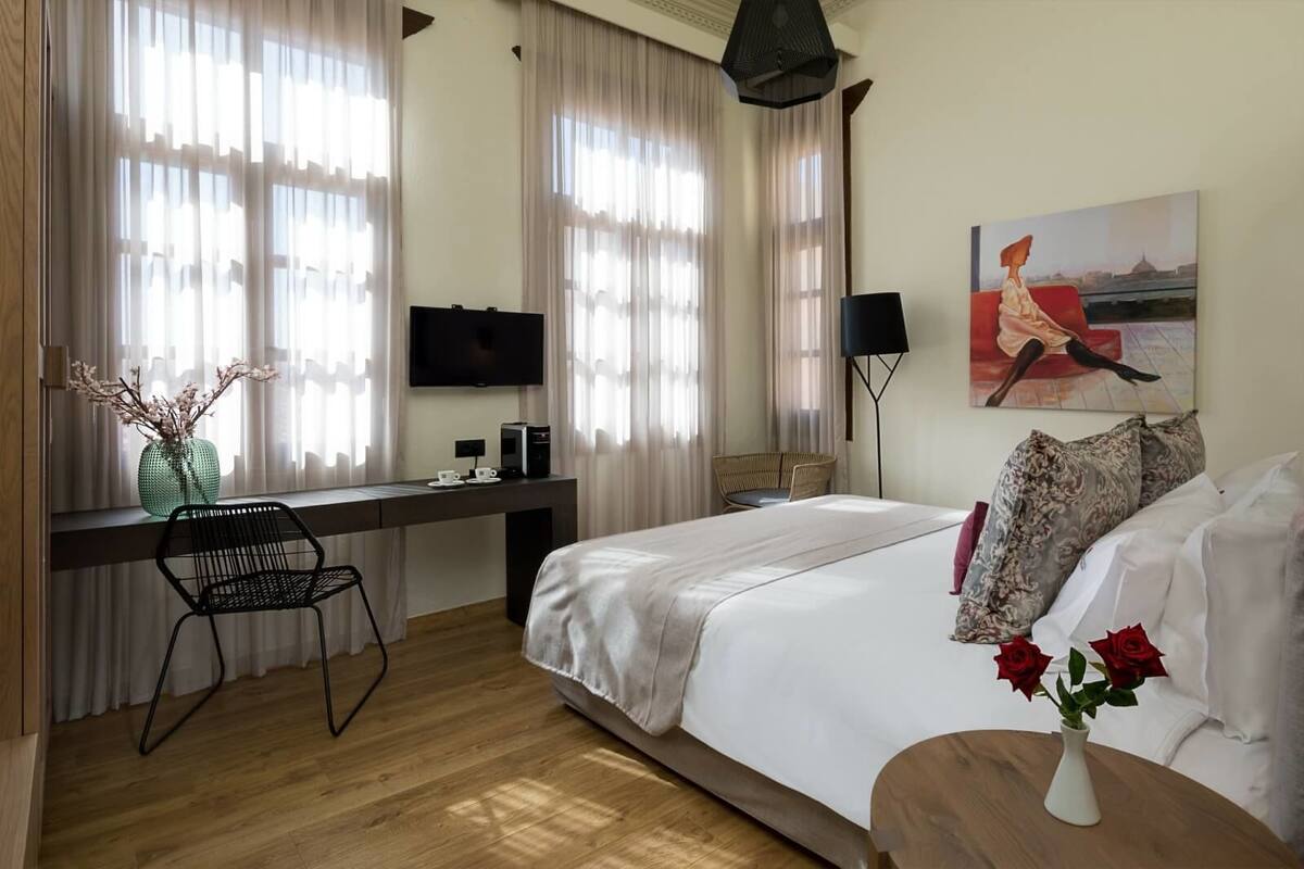 A hotel room with a bed, a desk with a computer, a chair, a TV, and a painting on the wall.