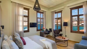 A hotel room with two beds, a desk, a chair, a small table, and large windows with a view of the sea and buildings. - Contessa Boutique Hotel (Chania)