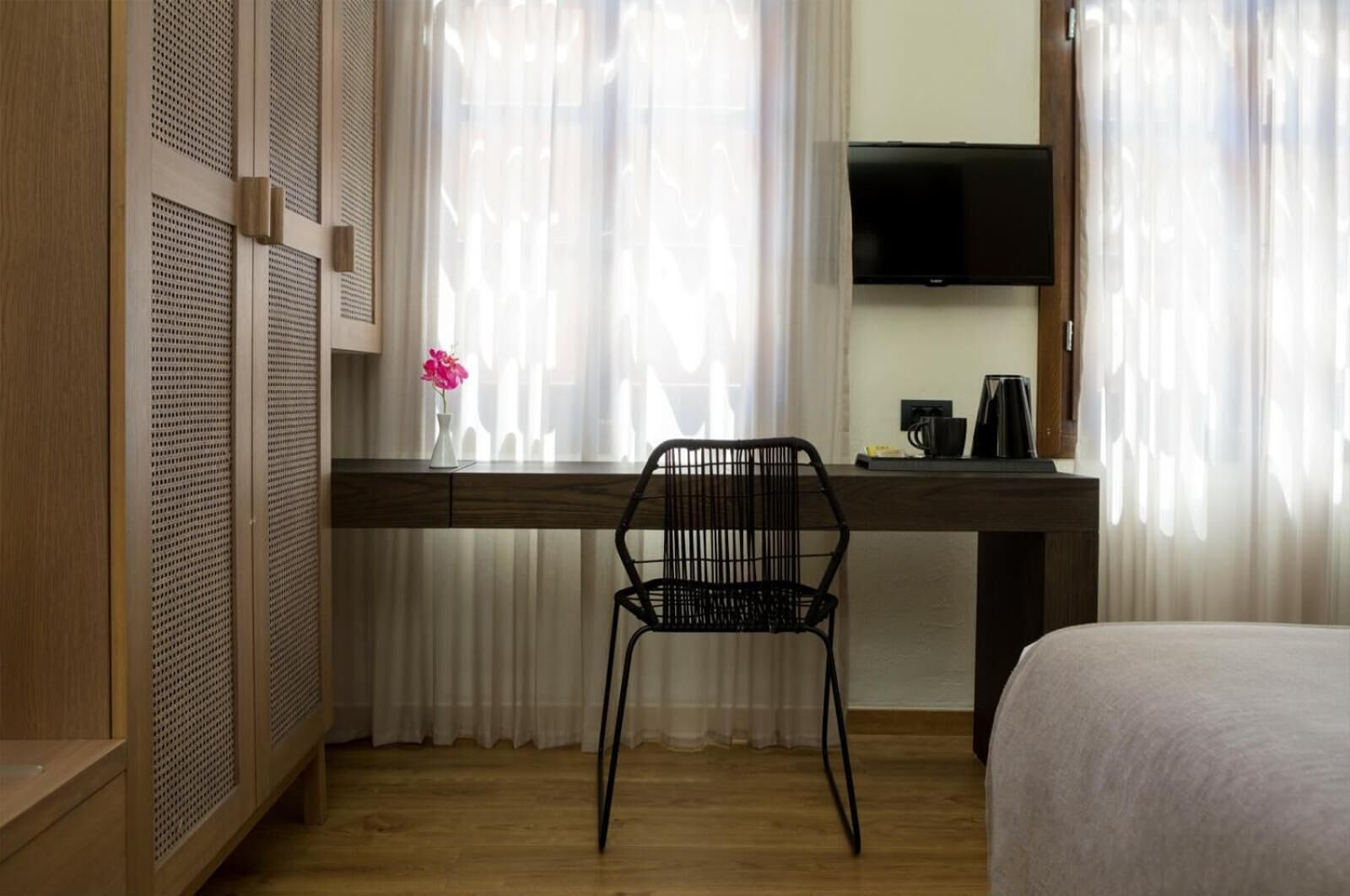 Habitación de hotel con un escritorio de madera, una silla negra, una cama, un televisor y una ventana con cortinas translúcidas.