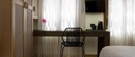 Quarto de hotel com uma escrivaninha de madeira, uma cadeira preta, uma cama, uma televisão e uma janela com cortinas translúcidas.