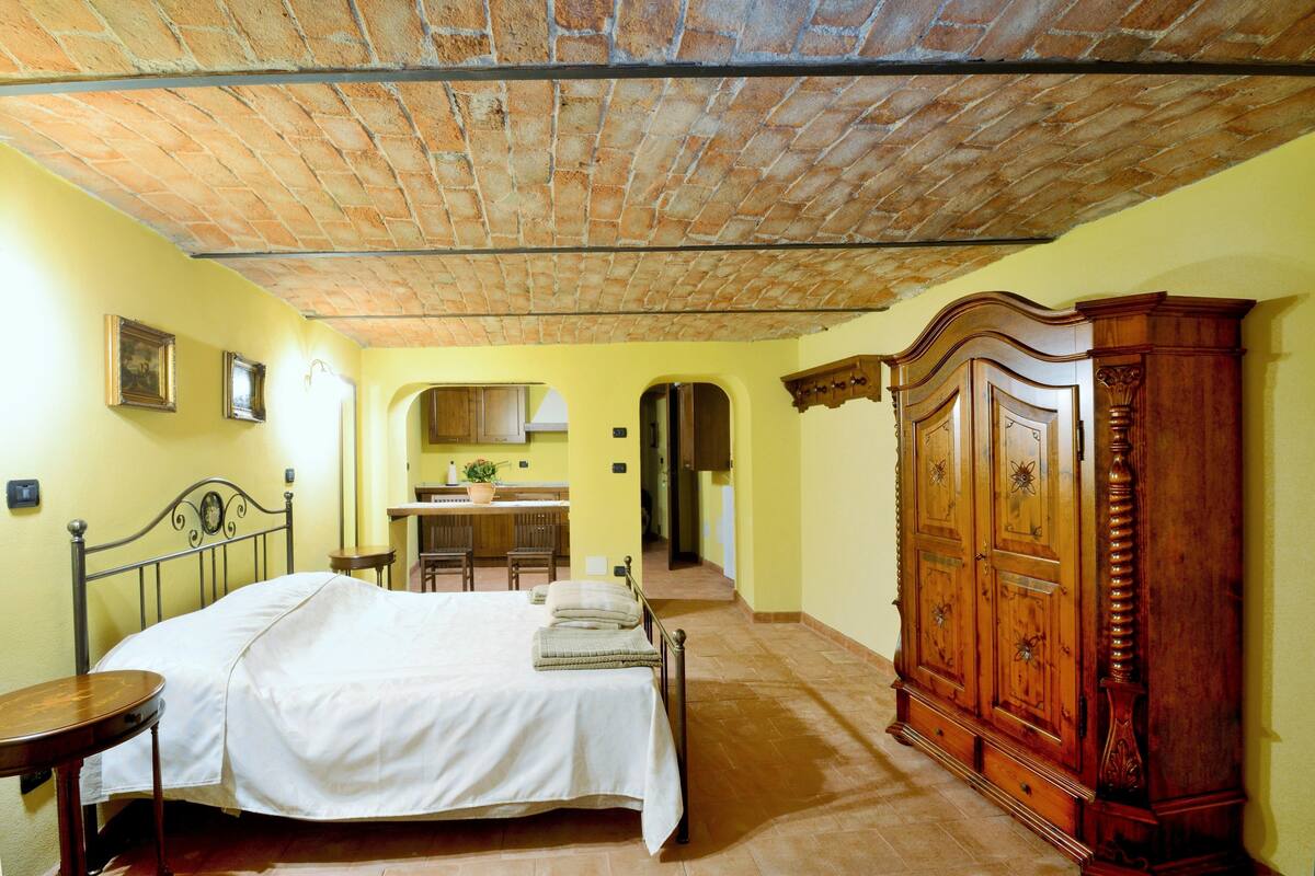 A bedroom with a bed, a wooden wardrobe, a desk with a chair, and a kitchen area in the background.