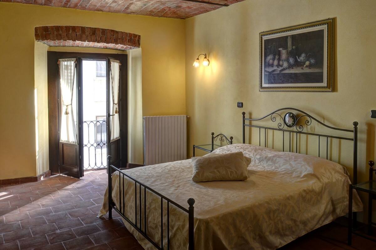 A bedroom with a brick ceiling, a bed with a white bedspread, a radiator, a framed picture on the wall, and a door leading to a balcony.