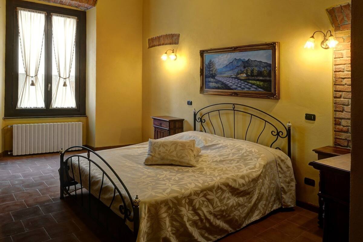 A bedroom with a bed, a window with curtains, a painting on the wall, and a chandelier.
