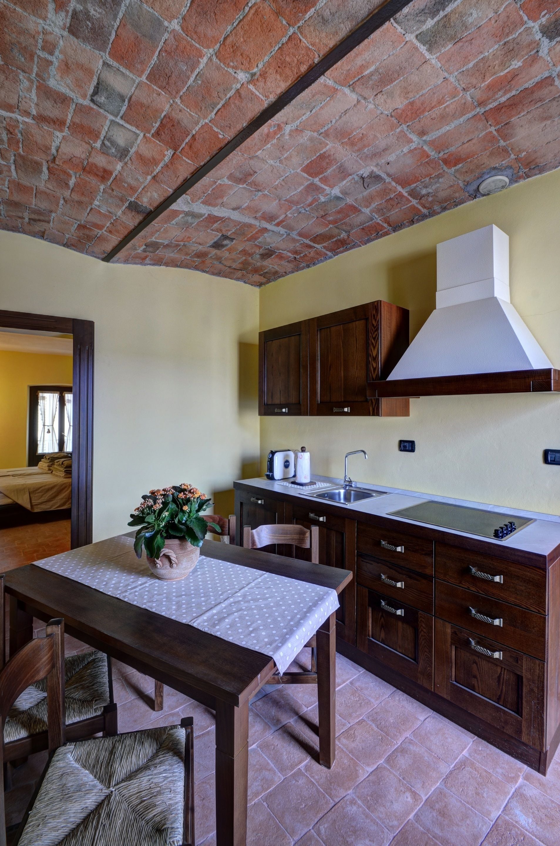 Una cucina con soffitto in mattoni, pensili in legno, cappa sopra il piano cottura bianca e un tavolo da pranzo con sedie.