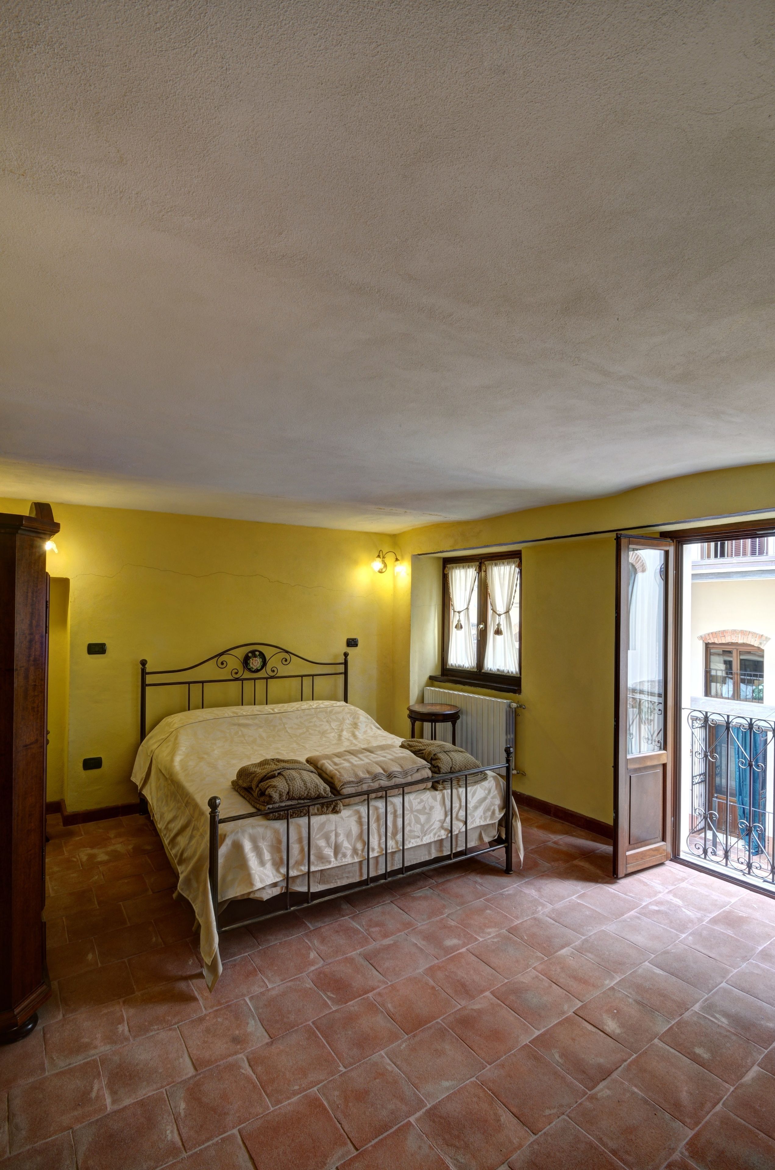 A bedroom with a bed, a nightstand, a window with curtains, and a balcony with a railing.