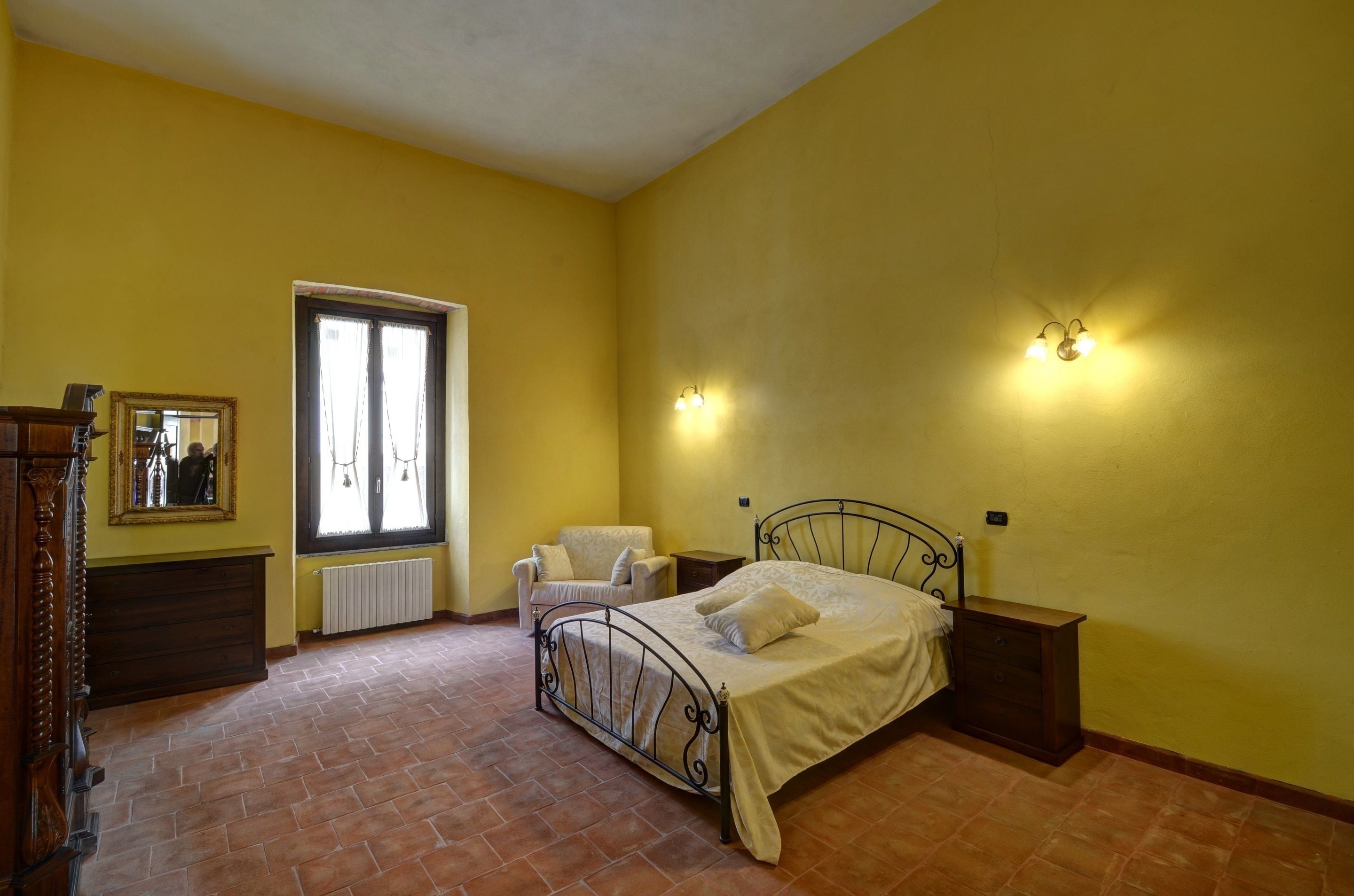 A bedroom with a bed, a chair, a dresser, a mirror, and two wall-mounted lights.