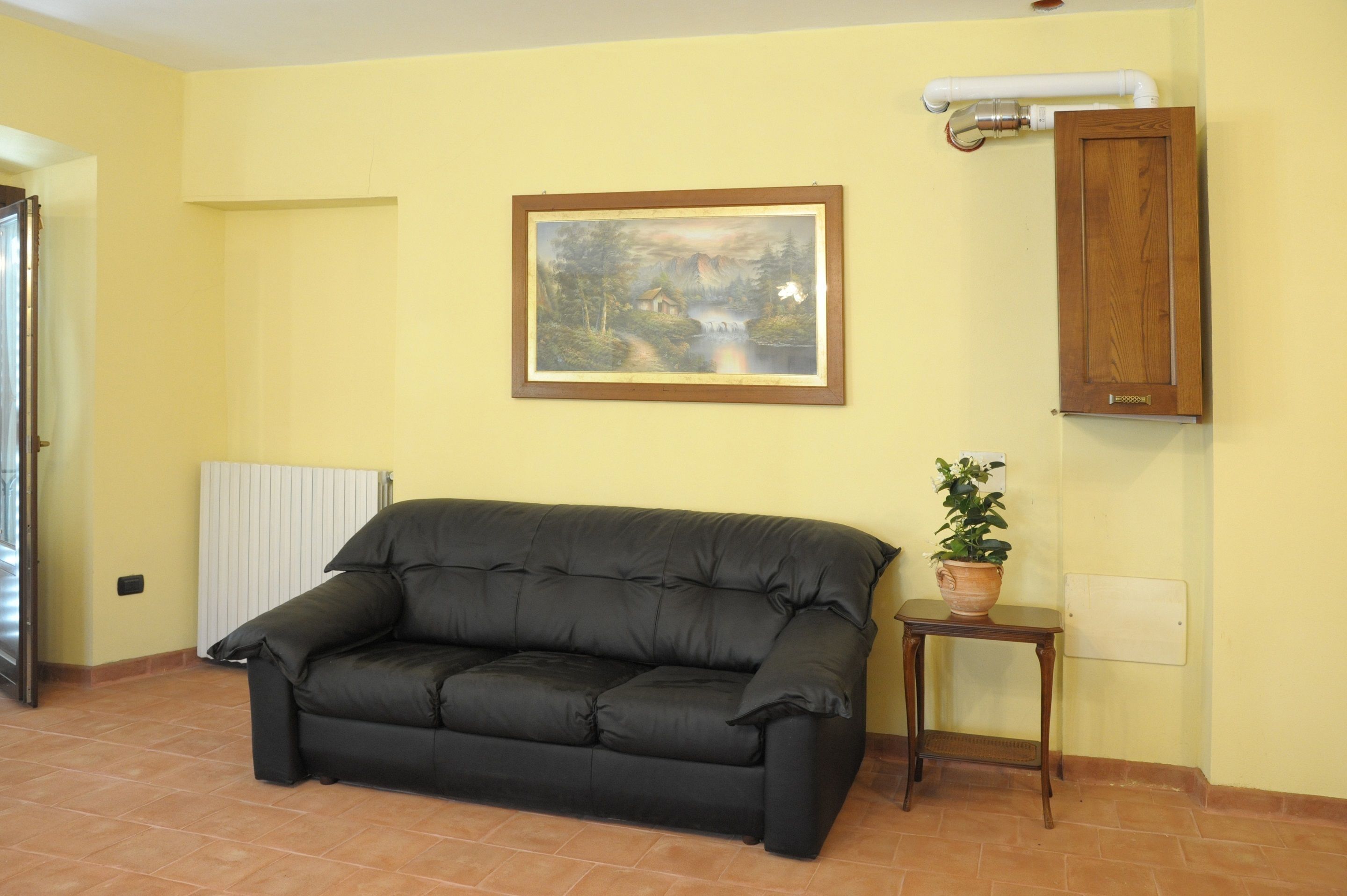 Un salón con un sofá negro, una mesita con una planta en maceta, un cuadro enmarcado y un mueble de madera.