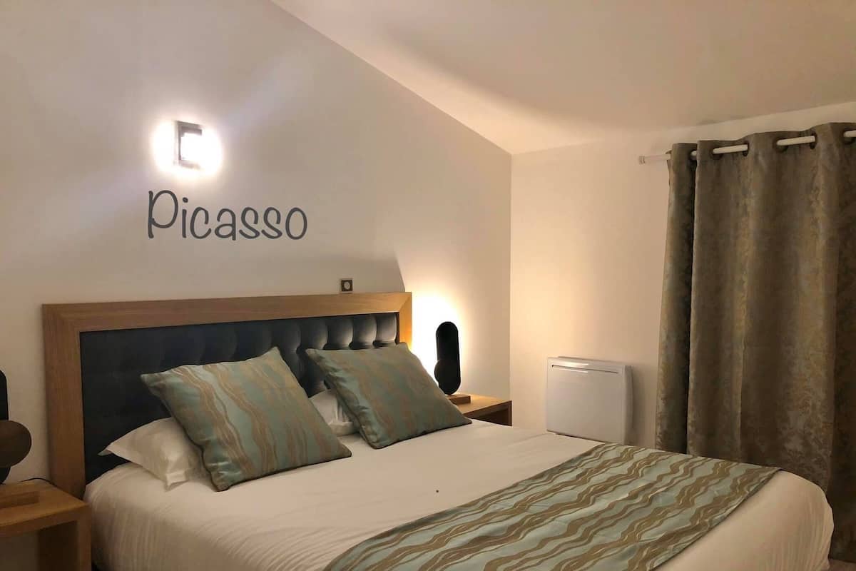 Um quarto com cama, mesa de cabeceira, luminária na parede, termostato e cortina.