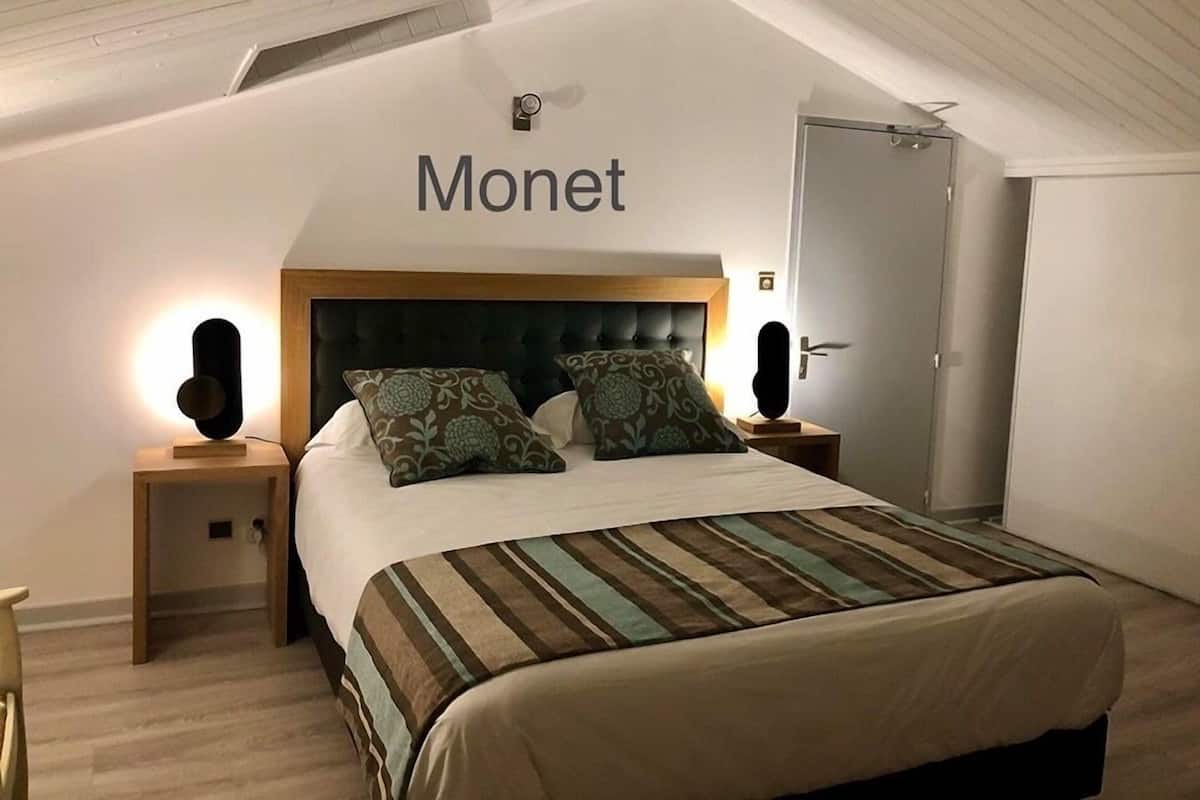 Quarto de hotel com cama, duas mesas de cabeceira, uma porta e teto com vigas aparentes.
