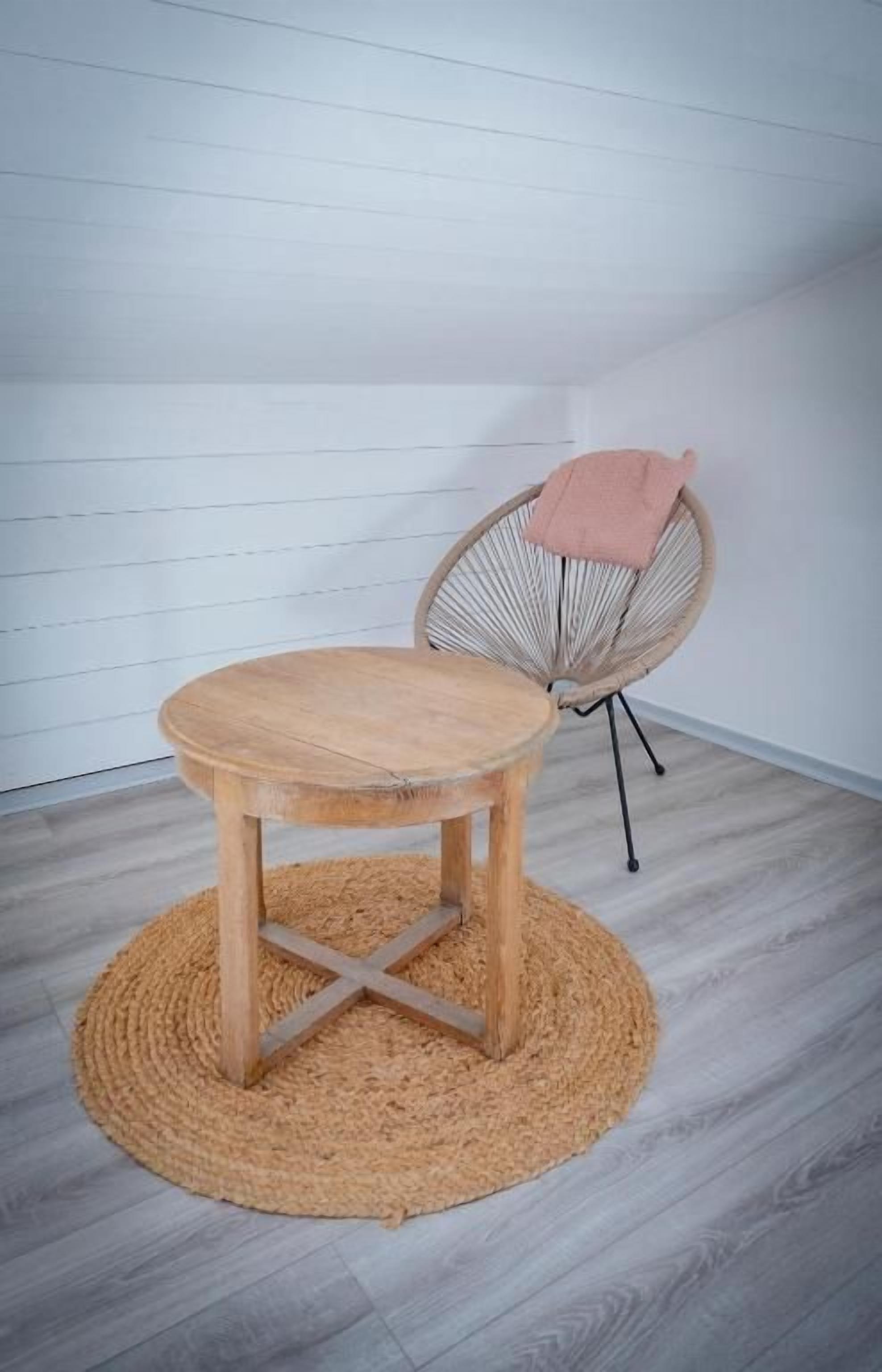 A round wooden table with a woven rug, a wicker chair with a pink towel, and a white wooden wall.
