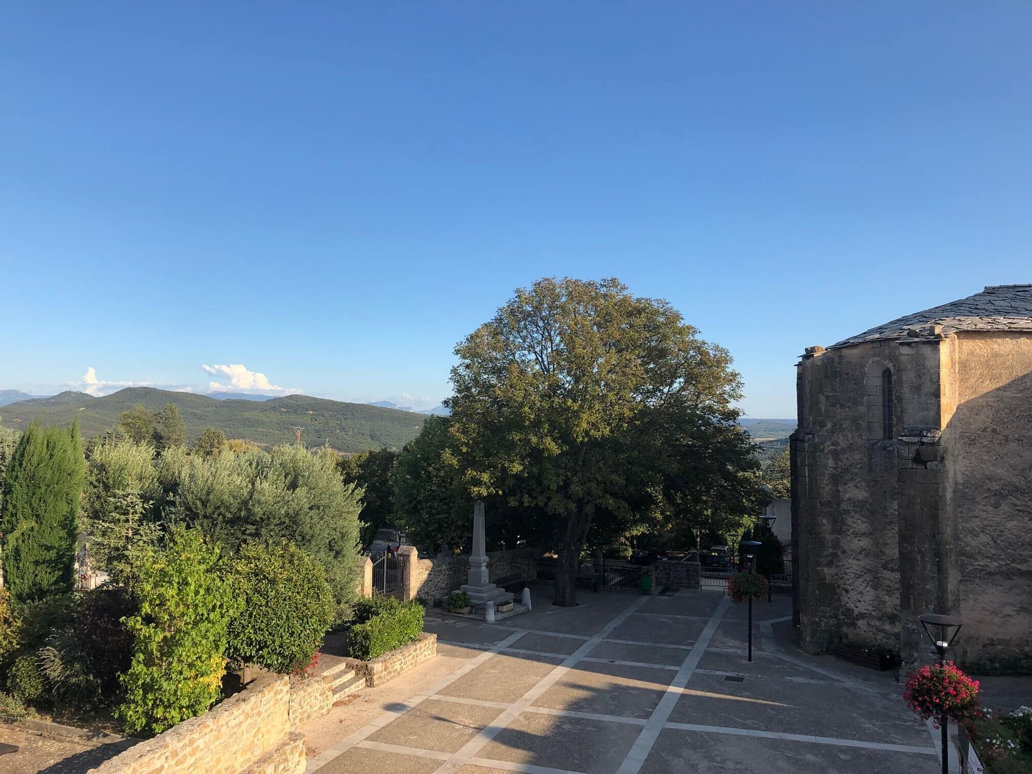 Un patio con un edificio de piedra, un árbol grande y vistas a colinas distantes.