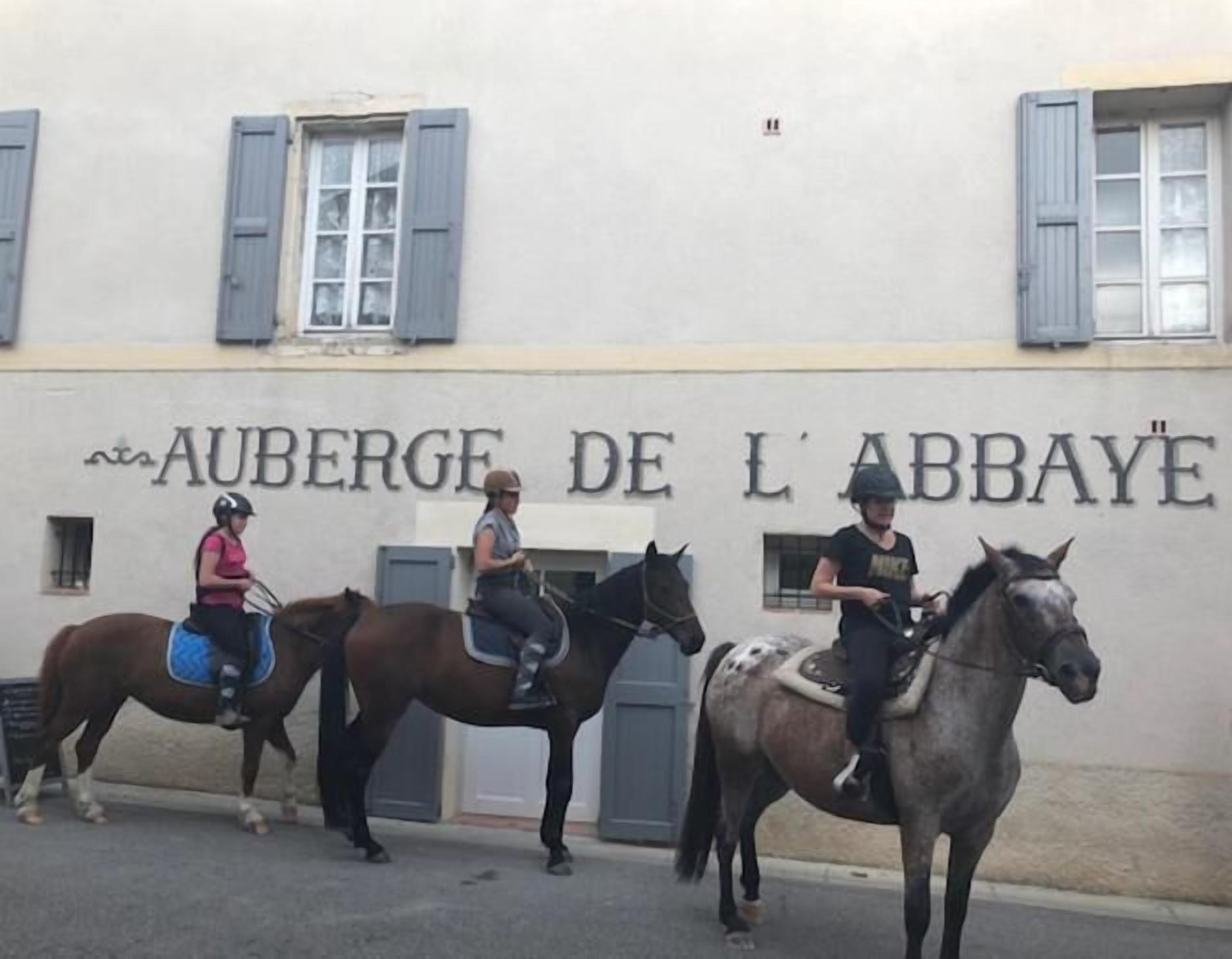 Trois personnes à cheval devant un bâtiment sur lequel on peut lire l’inscription « AUBERGE DE L’ABBAYE ».
