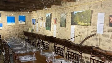 Una larga mesa de comedor con sillas, vigas de madera, paredes de piedra y obras de arte.