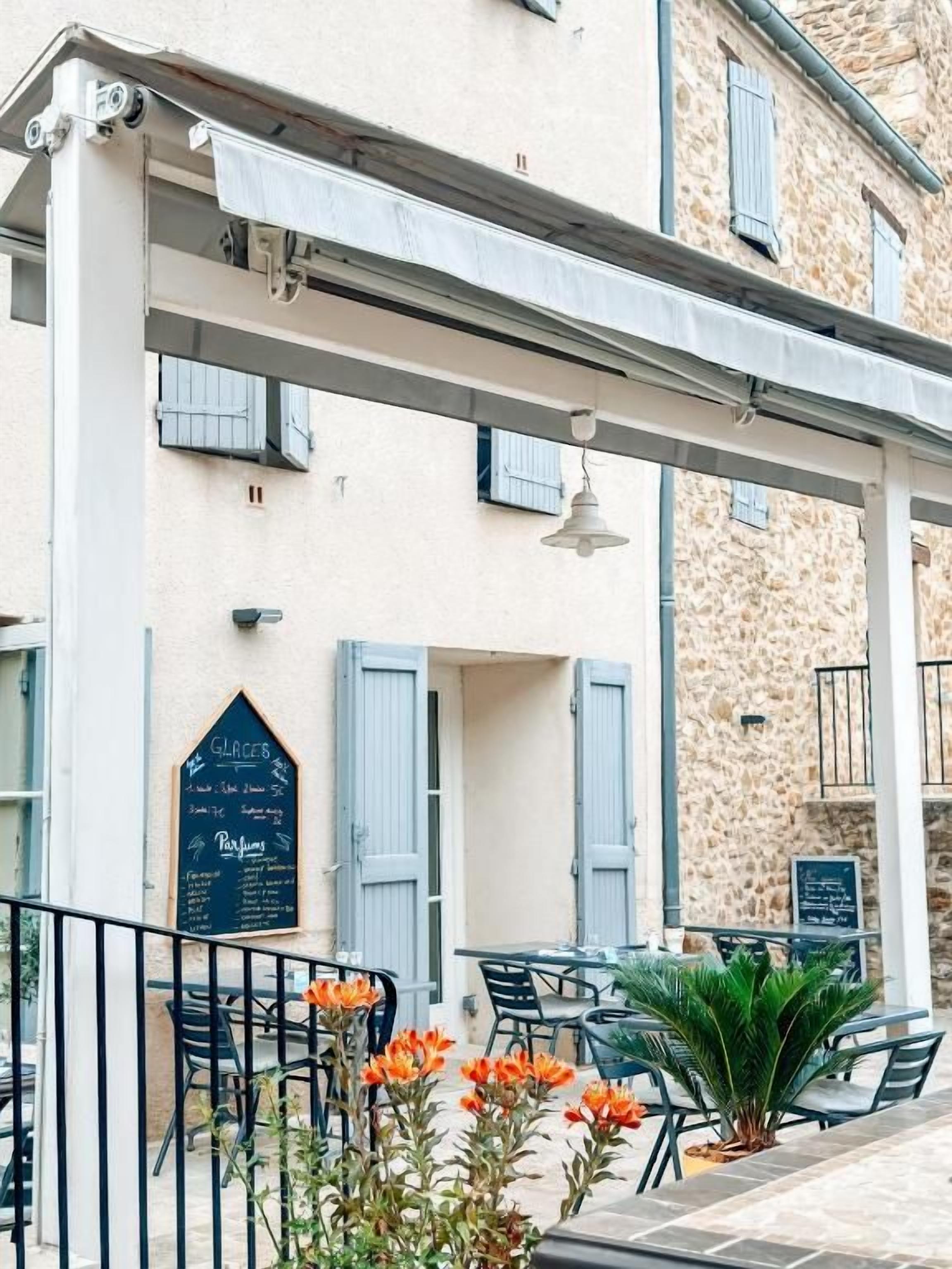An outdoor café with tables and chairs, a pergola, and a building with a stone facade.