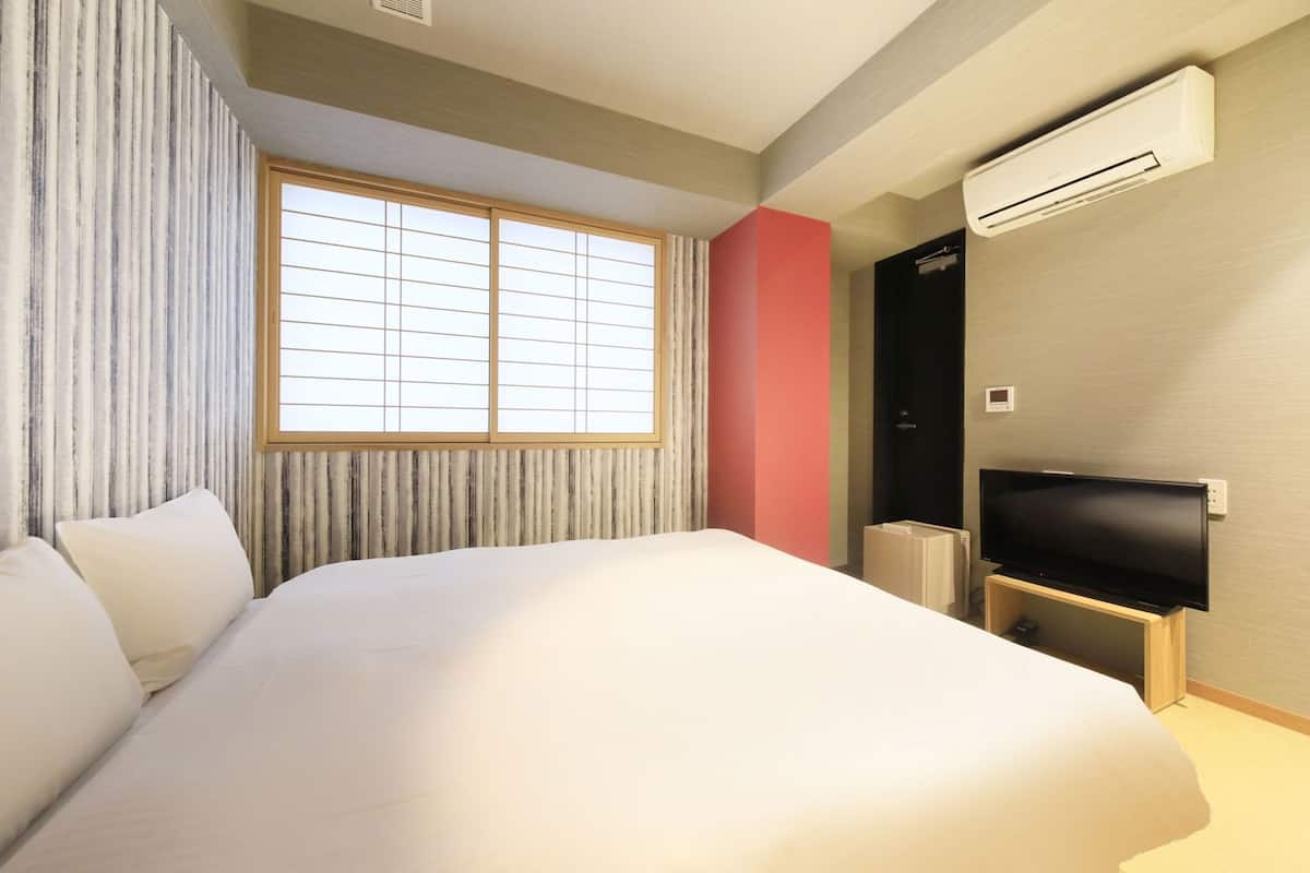 A hotel room with a large bed, a television, an air conditioning unit, and a window with blinds.
