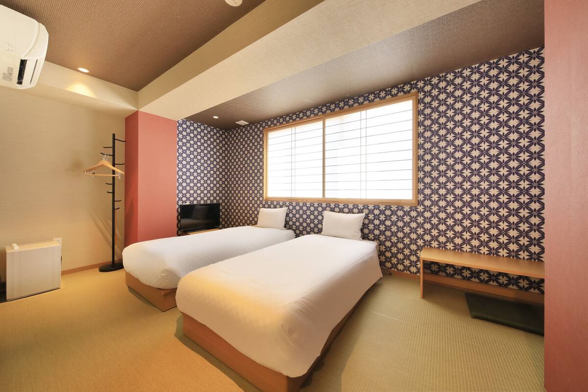 A hotel room with two beds, a large window with blinds, a TV, and a wardrobe.