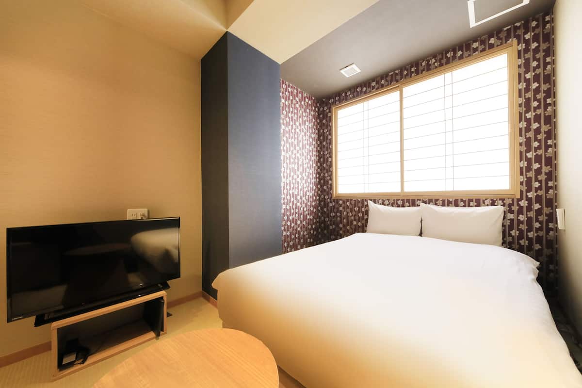 A hotel room with a bed, a TV on a stand, a window with blinds, and patterned wallpaper.