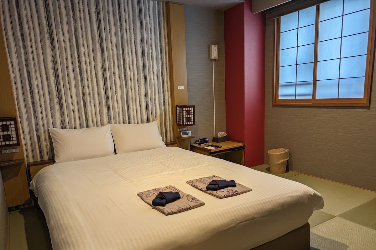 A hotel room with a large bed, a bedside table, a lamp, a trash bin, and a window with blinds.