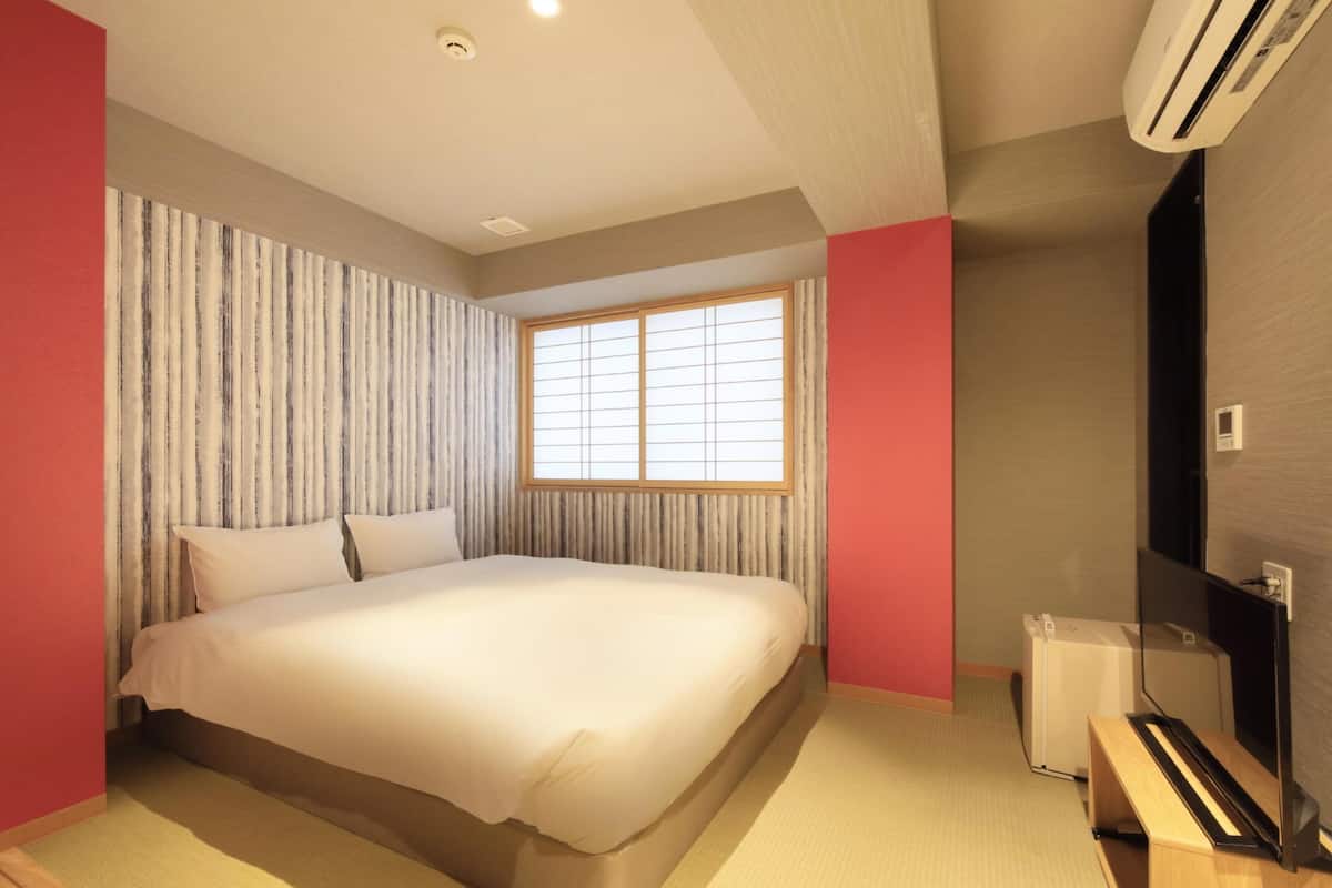 A modern bedroom with a large bed, a window with blinds, an air conditioning unit, and a wooden bench.