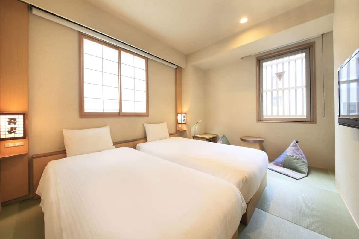 A hotel room with two beds, a small table, a chair, and a window with a view.