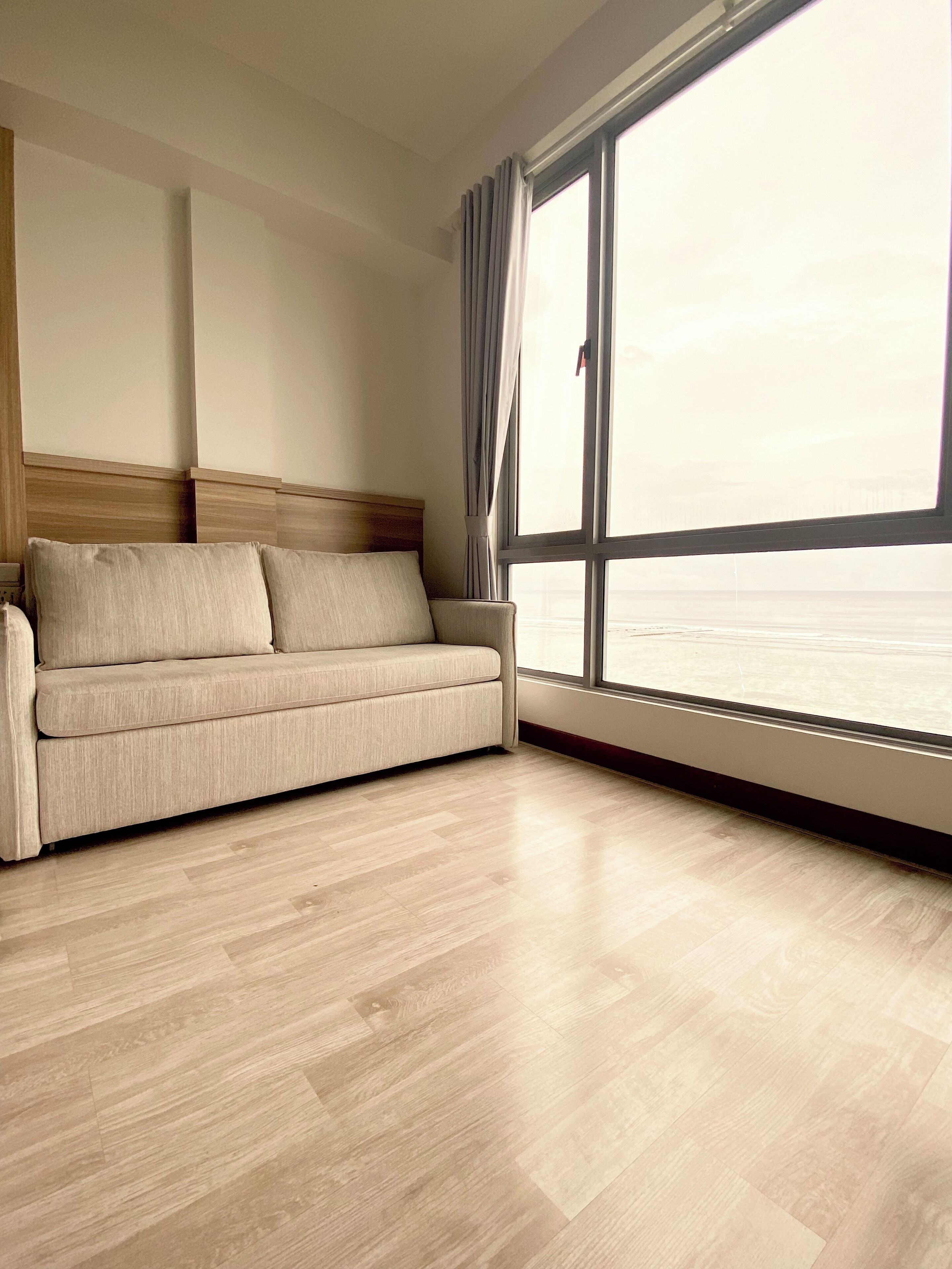 A room with a beige sofa, wooden flooring, and a large window with a view of the sea.