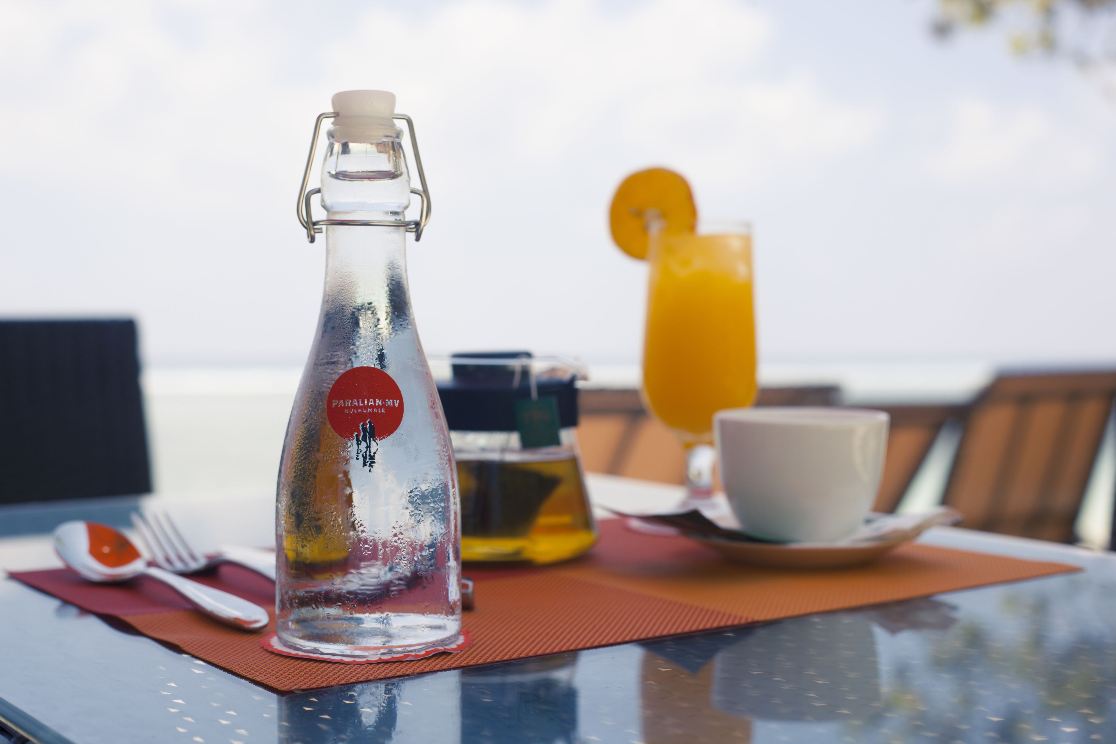 A table with a bottle, a glass of orange juice, a cup, and a fork on a glass table with a view of the ocean.