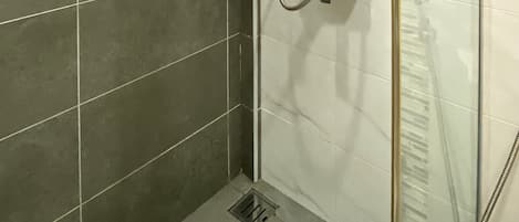 Zona de ducha con puerta de vidrio, cabezal de ducha manual y grifo manual. Hay un estante pequeño con una botella de champú y una de jabón corporal.