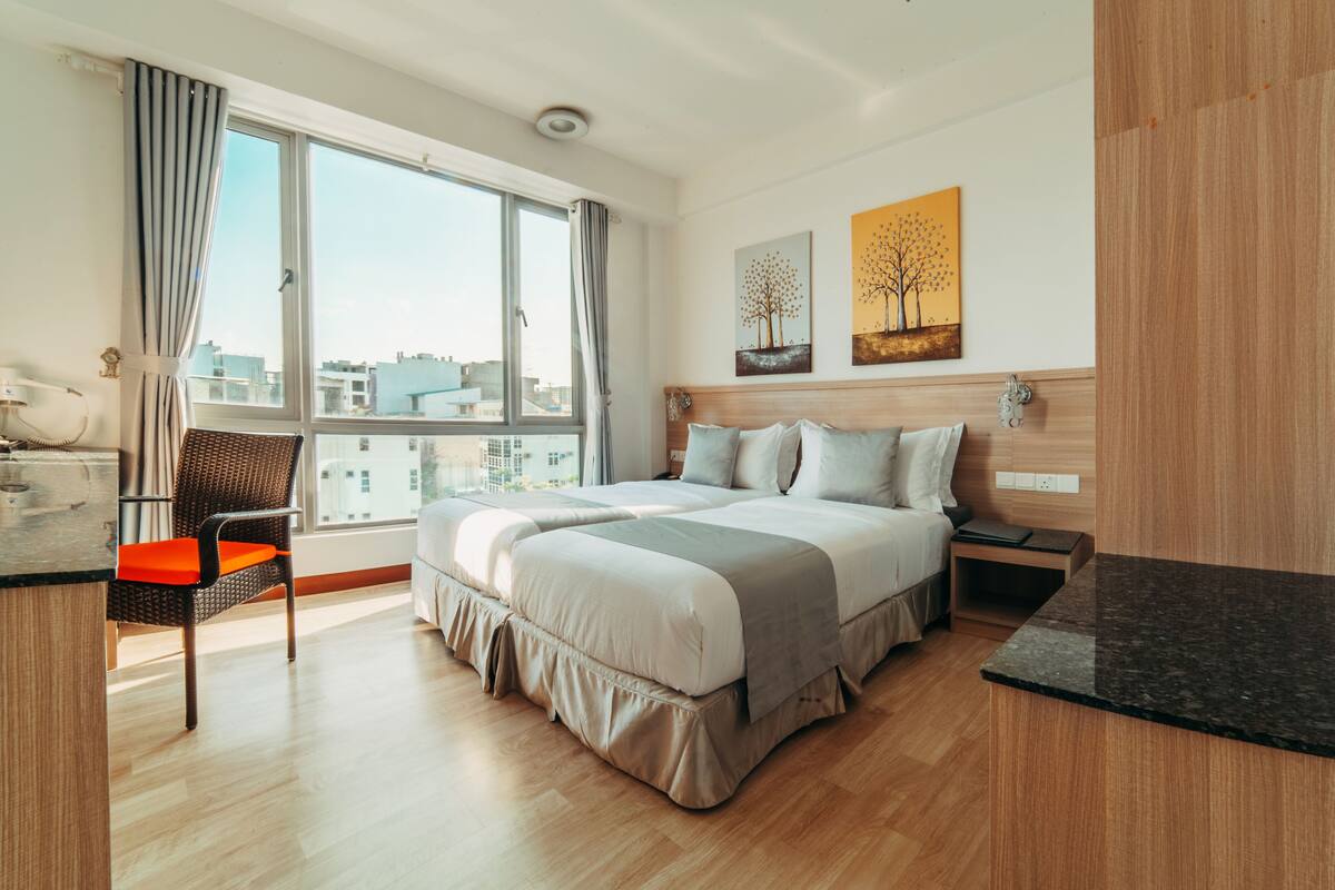 A modern hotel room with a large bed, a desk, and a chair. There is a window with a view of buildings outside.