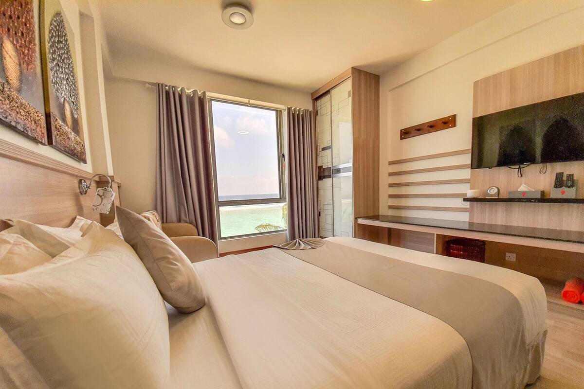 A hotel room with a large bed, a TV, a desk, and a view of the ocean.