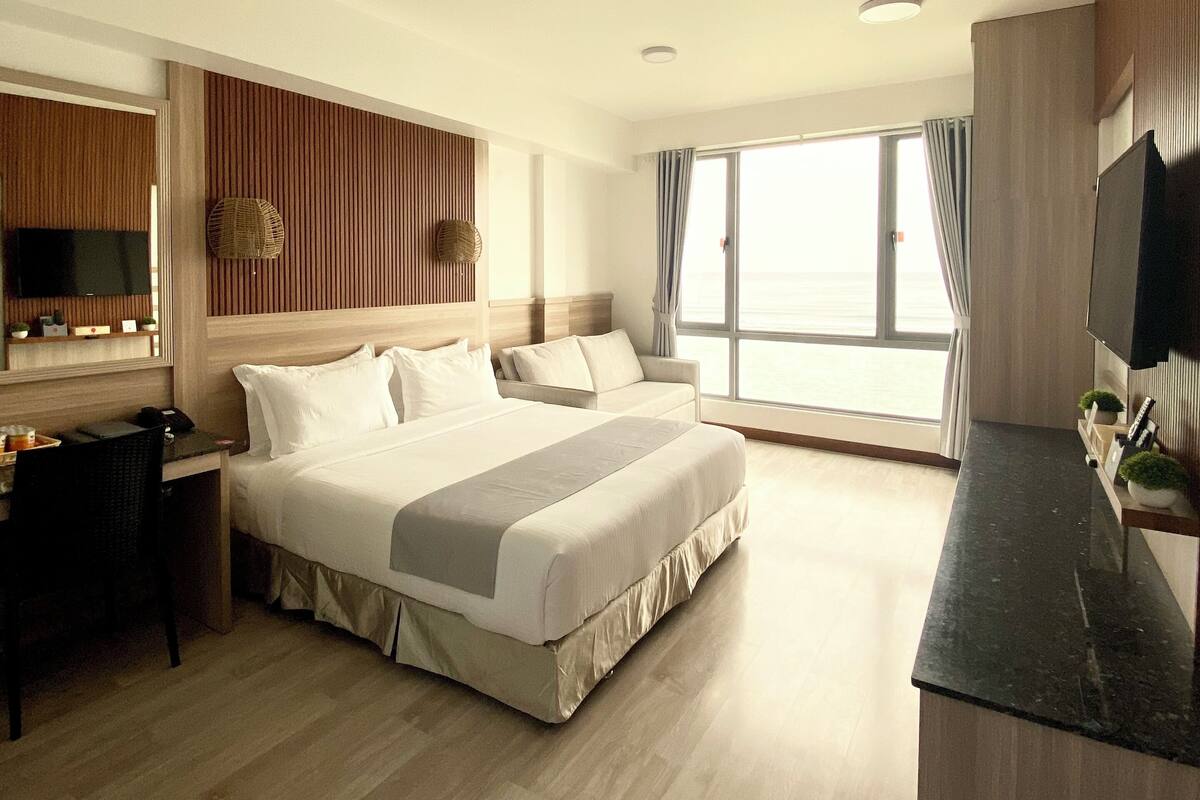 A hotel room with a large bed, a desk with a chair, a TV, and a view of the sea through large windows.