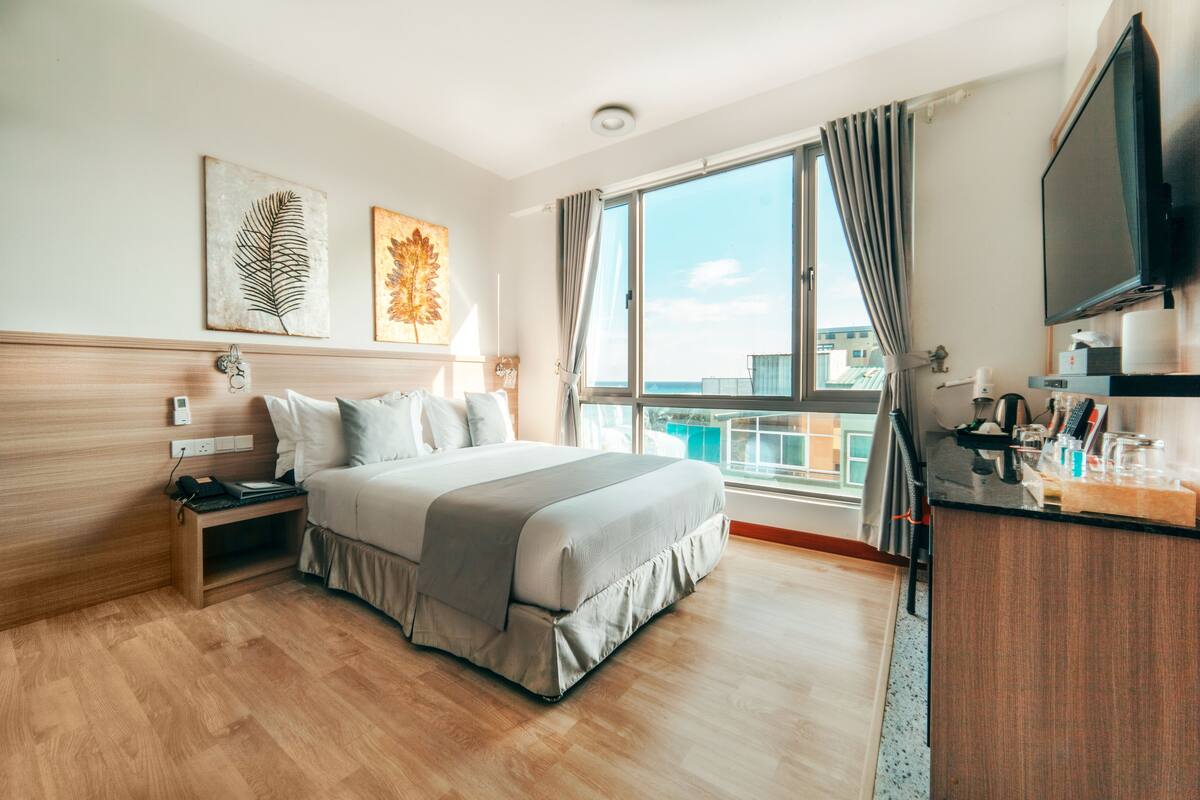A hotel room with a large bed, a desk with a flat-screen TV, and a balcony with a view of the city.