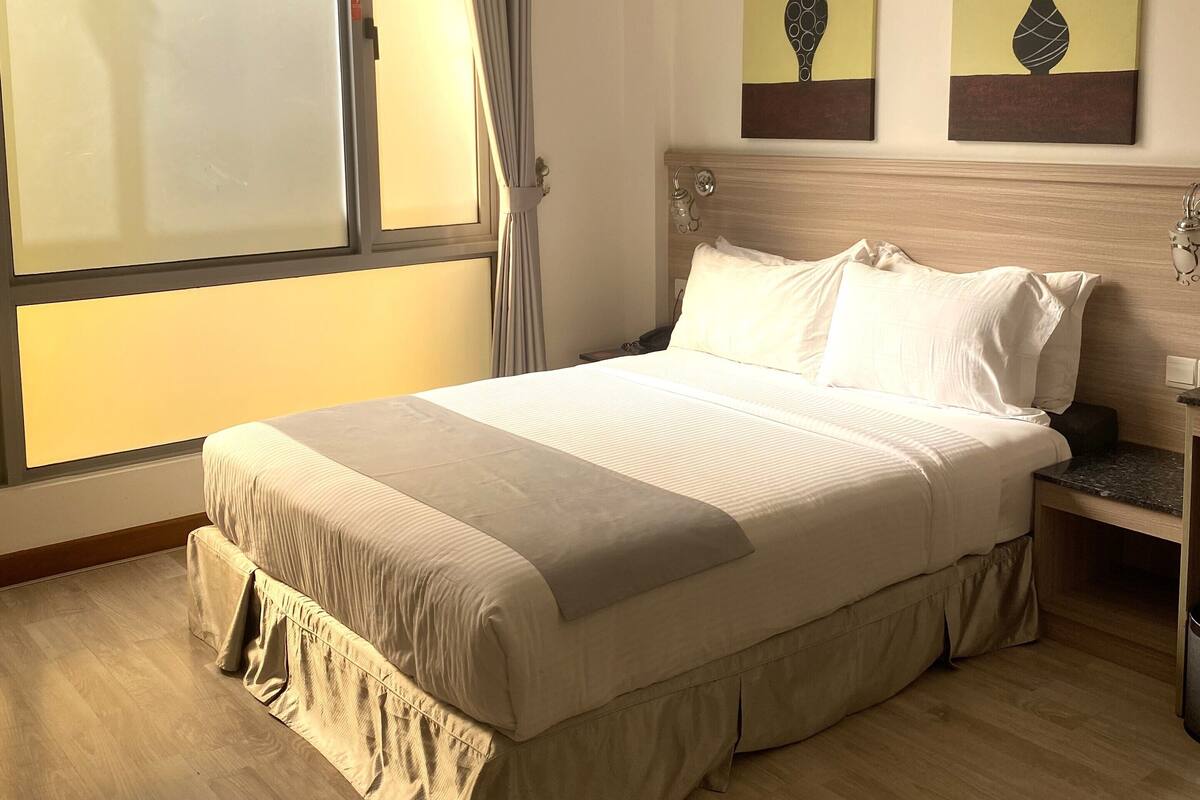 A hotel room with a large bed, a nightstand, a window with curtains, and two wall-mounted paintings.