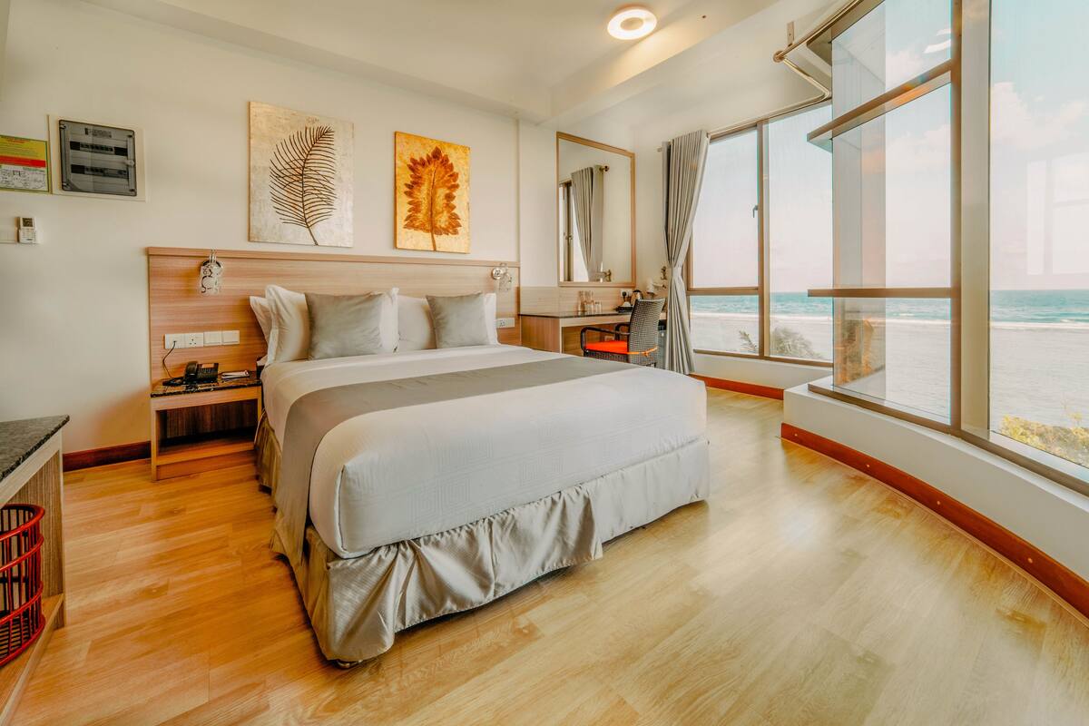 A modern hotel room with a large bed, a desk, and a view of the beach.