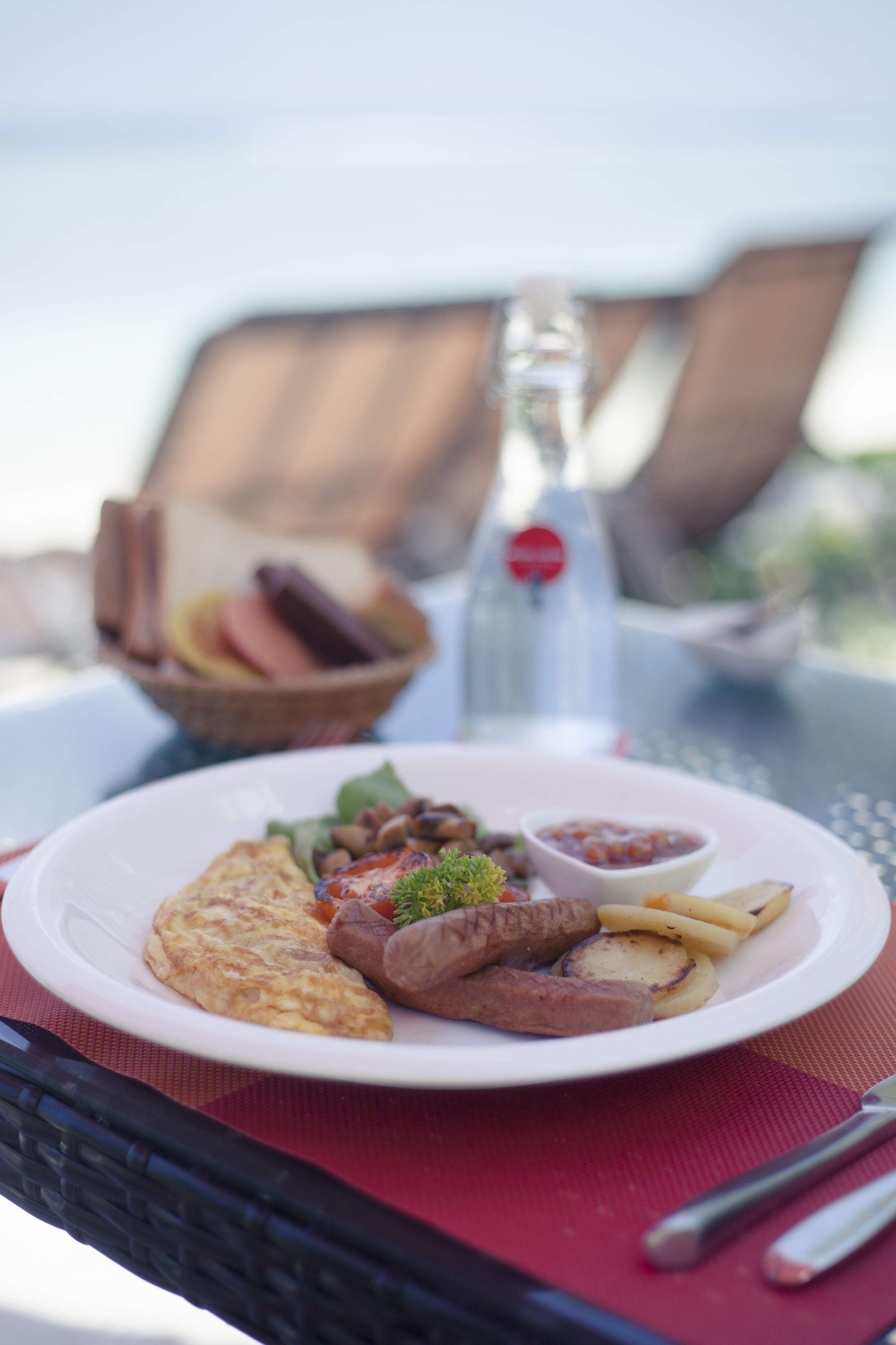 A plate of food with sausages, a slice of pizza, and a small bowl of sauce on a table with a red placemat.