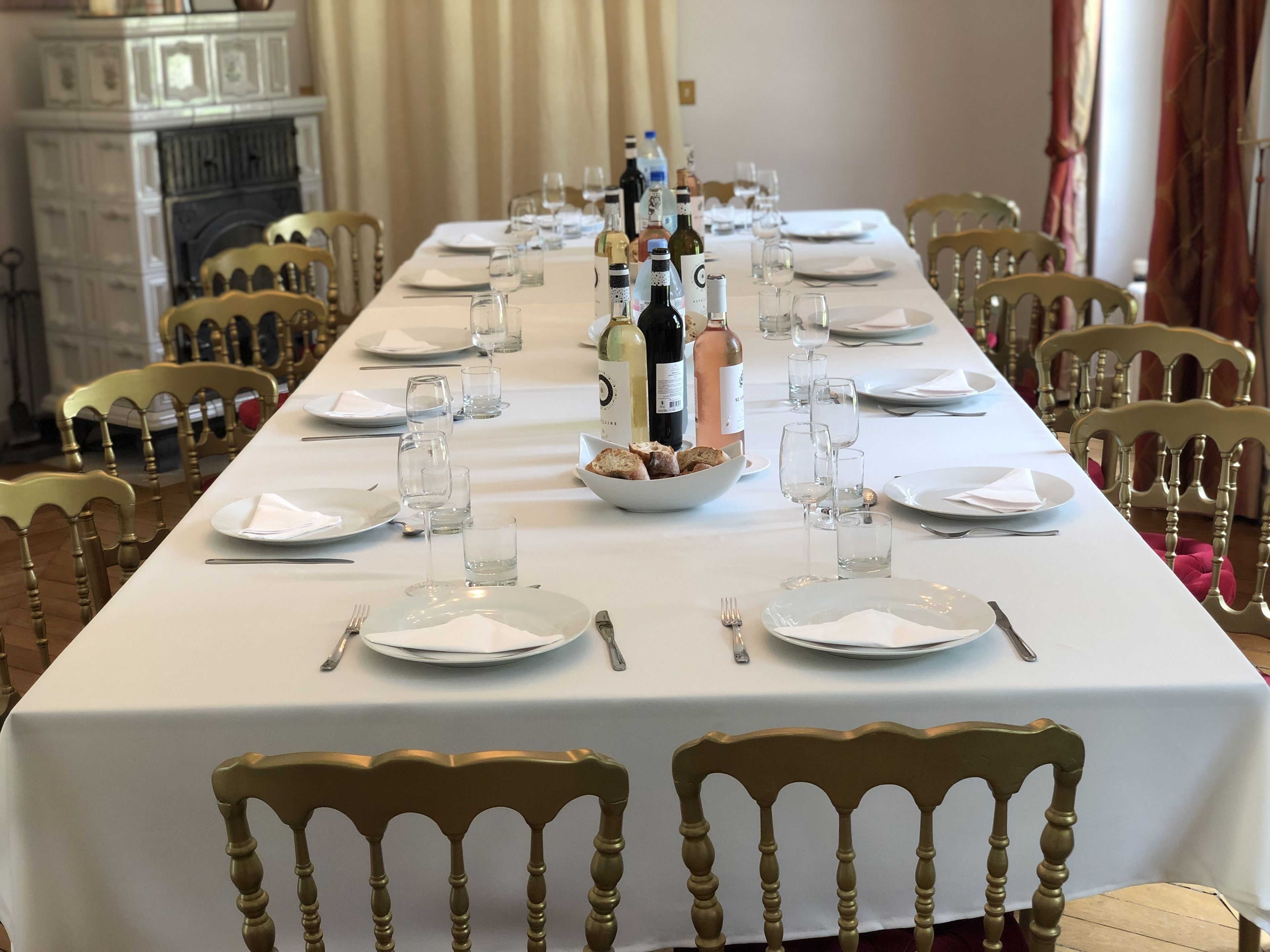 Una larga mesa de comedor con mantel blanco, platos, vasos y botellas, rodeada de sillas de madera.