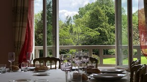 Mesa de comedor para cuatro personas, con copas de vino, platos y pan, con vistas a un paisaje verde exuberante a través de amplios ventanales.