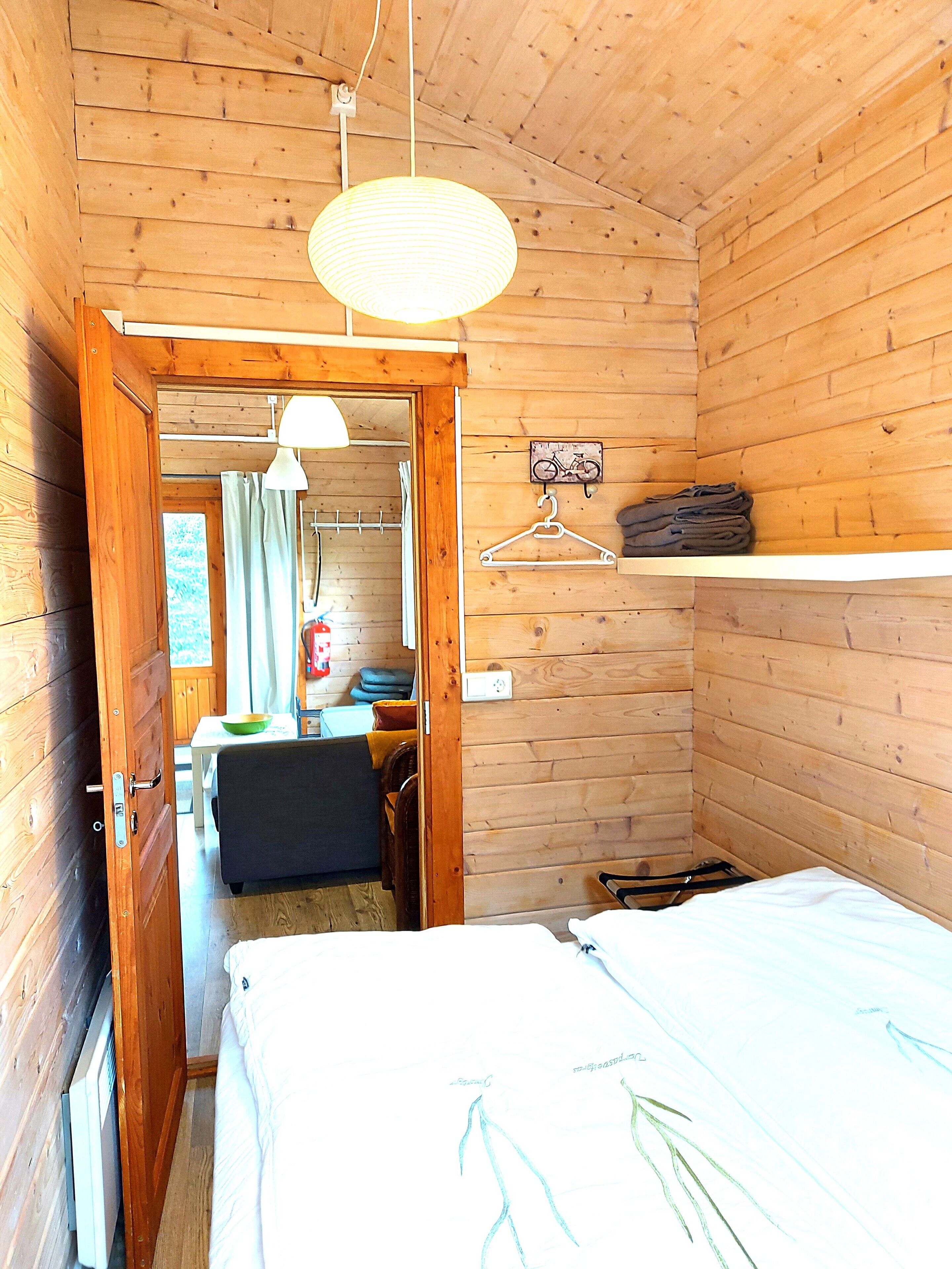 Una habitación de cabaña de madera con una cama, una lámpara colgante, una mesita con una silla y una bicicleta como adorno en la pared.