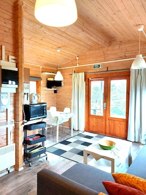 A wooden cabin interior with a dining area, kitchenette, and a large window. - Ásgeirsstaðir Cottages (Eidar)